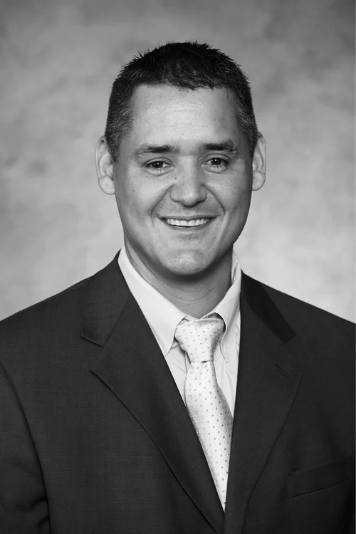 A smiling man in a dark suit with a light shirt and a patterned tie standing against a neutral background.