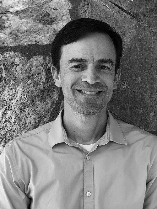 Close-up photo of a man smiling, wearing a light blue button-up shirt, standing in front of a textured stone wall with varying shades of brown, beige, and gray.