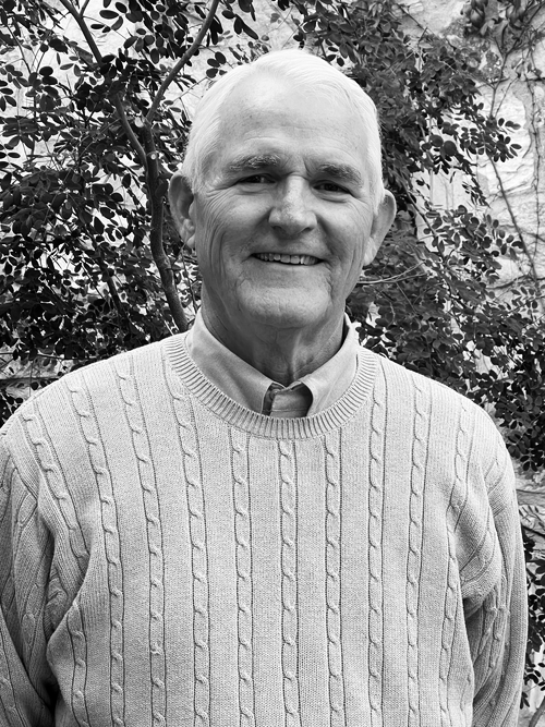 A smiling elderly man with white hair wearing a light gray cable knit sweater and a collared shirt, standing outdoors with green foliage and a brick wall in the background.