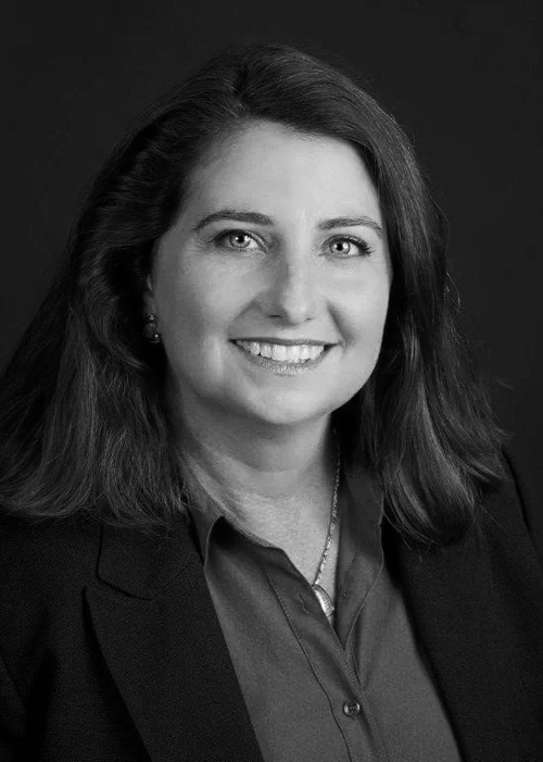 Professional portrait of a woman with shoulder-length brown hair, wearing a navy blazer and a blue blouse, smiling against a dark background.