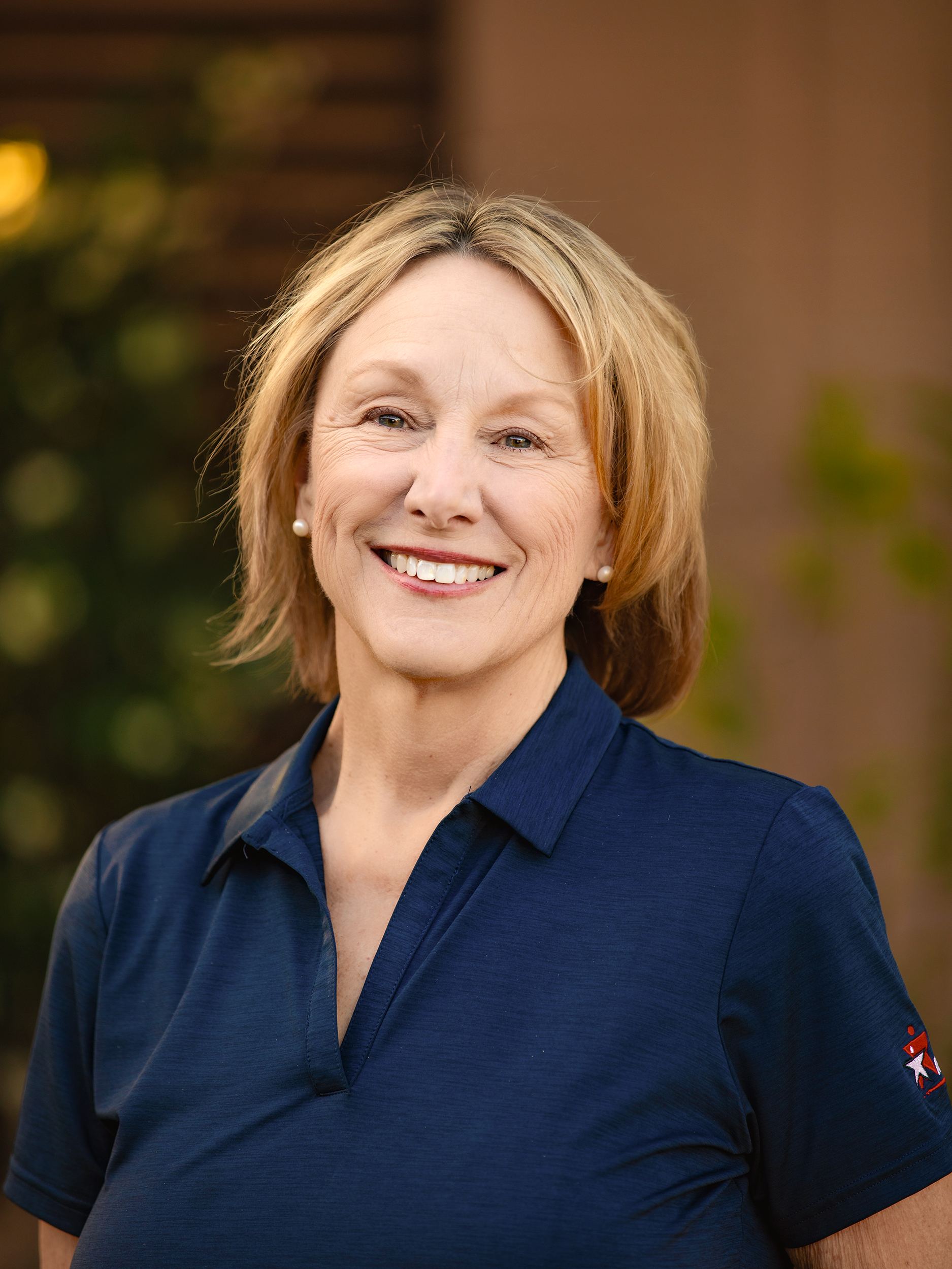 Close-up of a smiling woman with blonde hair wearing a navy blue shirt with a Texas flag patch on the sleeve, standing outdoors with blurred background.