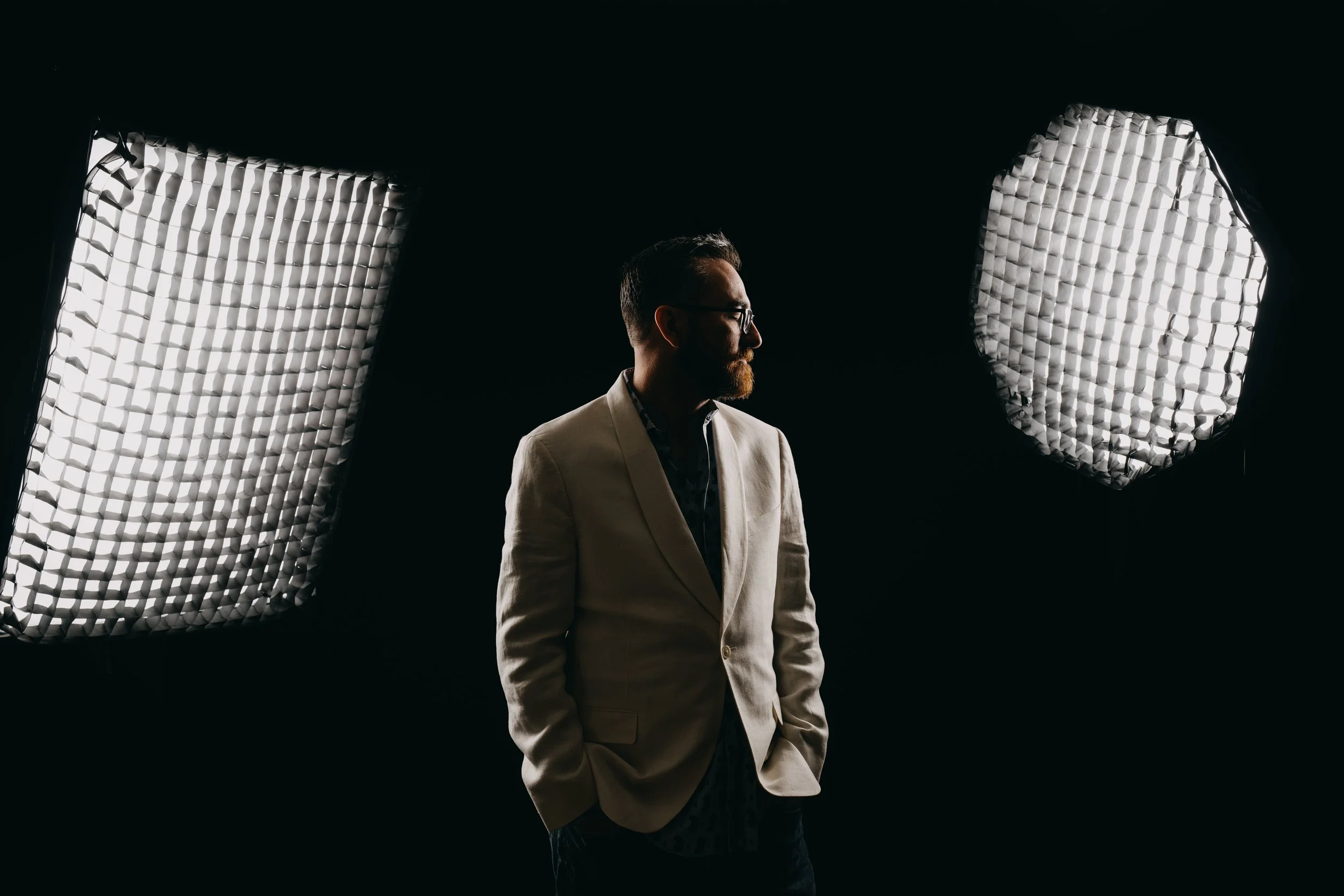 A man with a beard, wearing glasses and a light-colored blazer, standing in a dark room surrounded by two large studio lights.