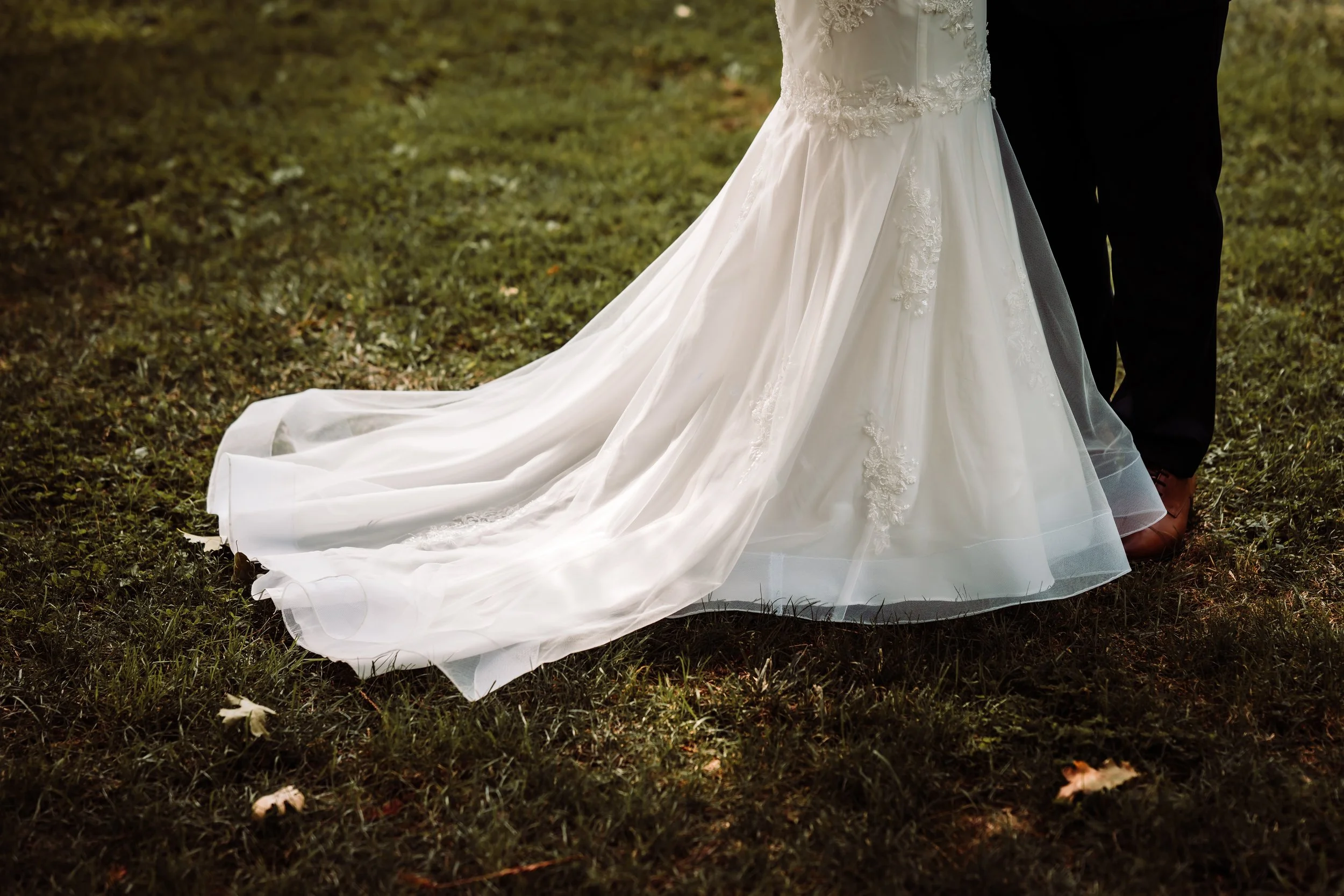 Close-up of a bride in a white wedding gown and a groom in black suit, standing on grass outdoors, with the focus on their lower bodies and the bride's long train.