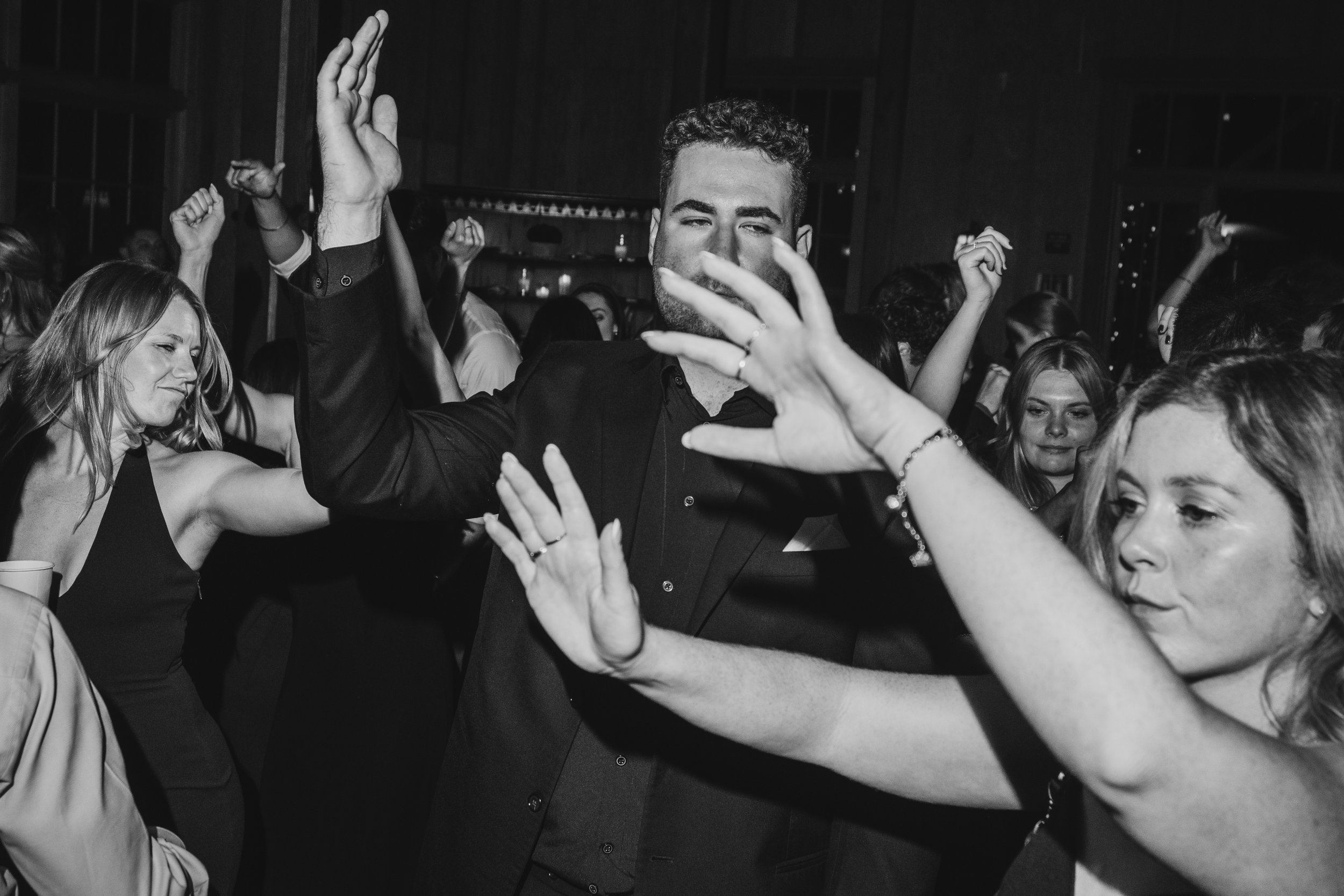 A black and white photo of a man with curly hair and a beard dancing at a crowded party or nightclub, with people around him also dancing or enjoying themselves.