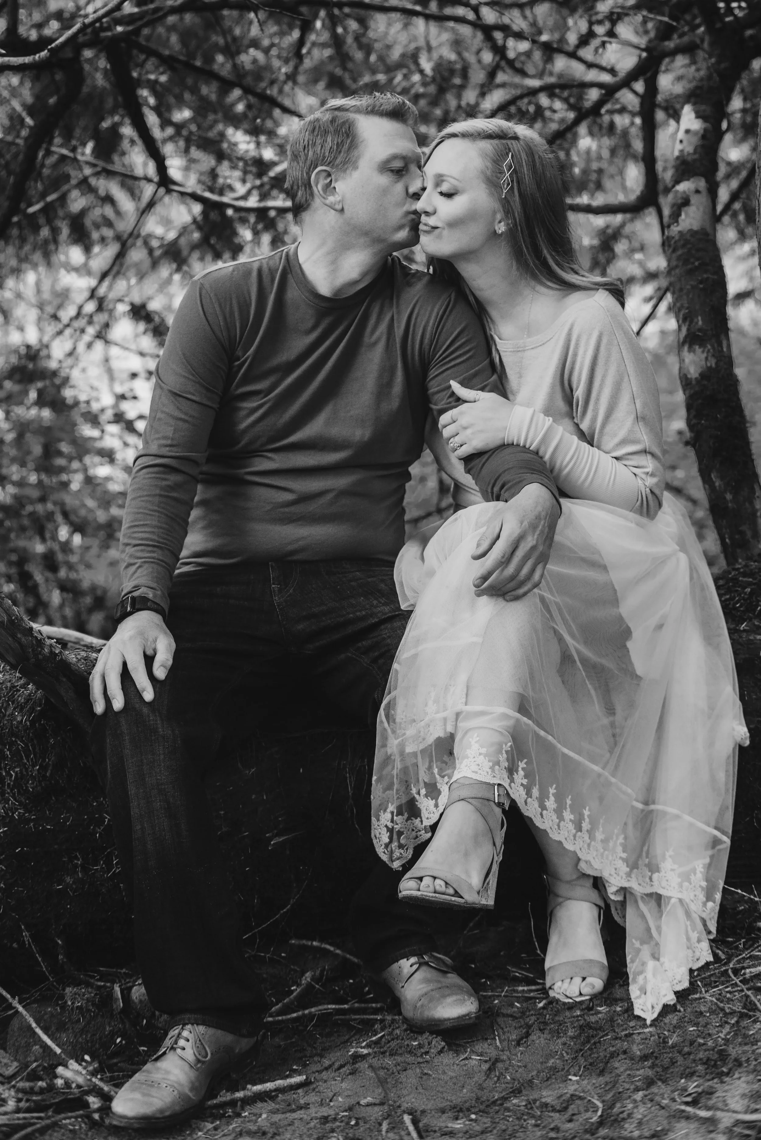 A black and white photo of a man and woman sitting together outdoors among trees. The man is kissing the woman on the forehead while she blissfully closes her eyes. The woman is wearing a light dress with lace details, and high-heeled shoes. The man is in a dark long-sleeved shirt and jeans. They are sitting on a tree branch or similar natural setting.