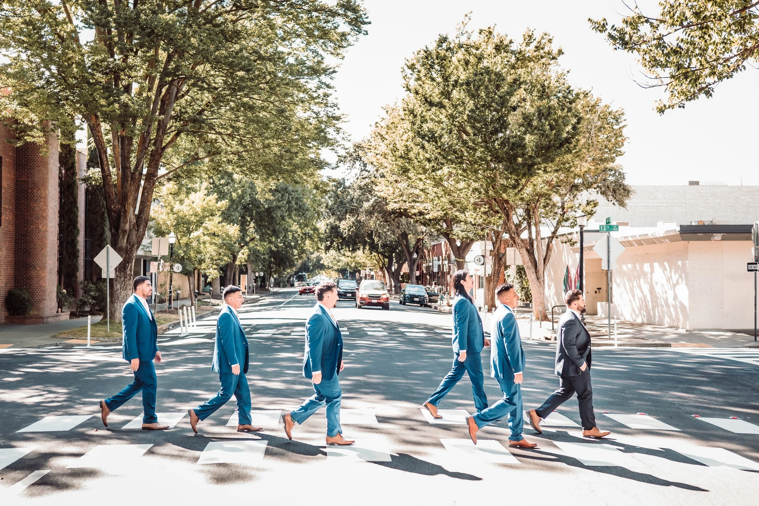 A group of seven people in blue suits walking across a crosswalk on a city street lined with trees and buildings, with cars parked along the sides.