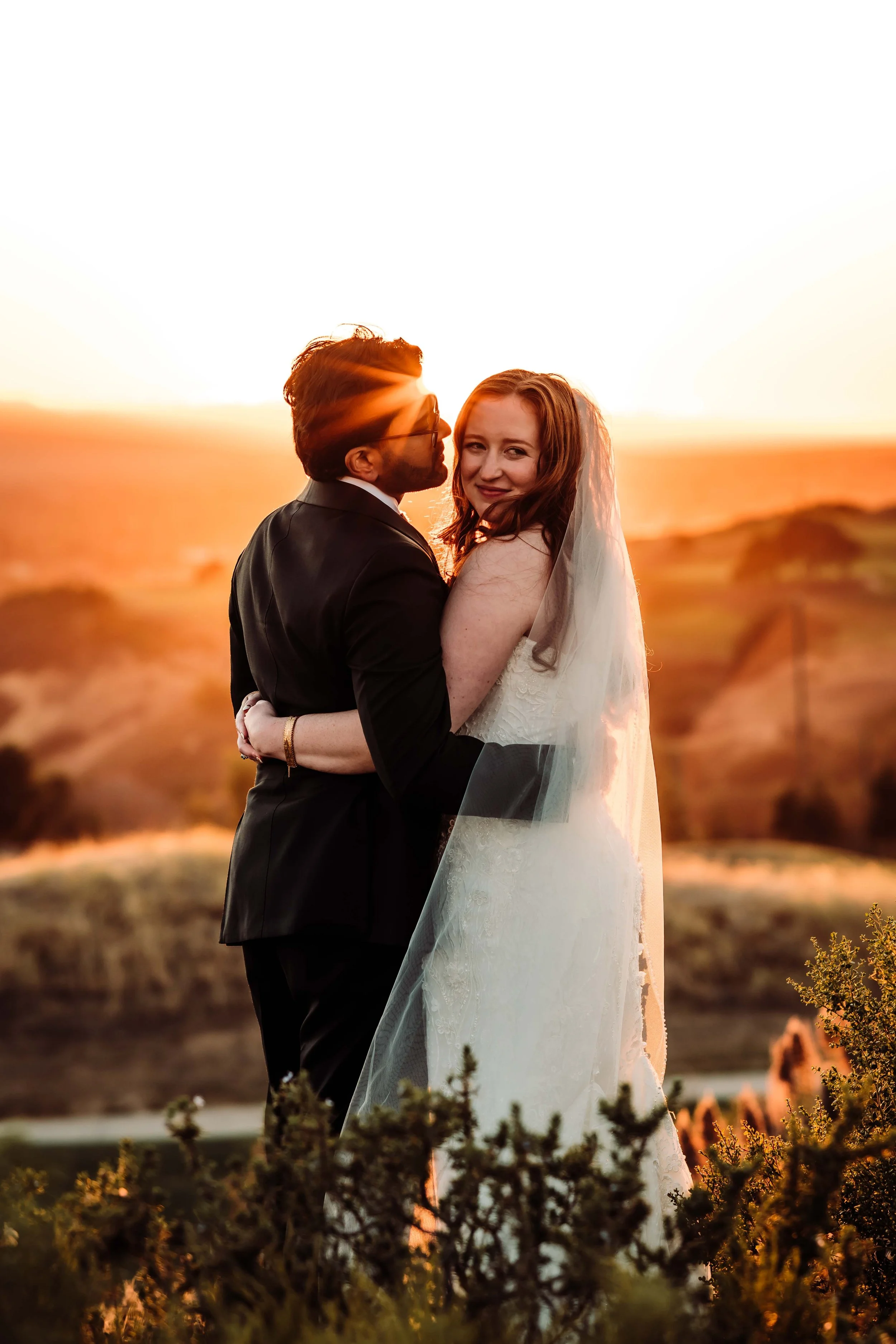 A couple in wedding attire embracing during sunset, with the woman in a white wedding dress and veil, and the man in a black tuxedo, on a scenic outdoor landscape.