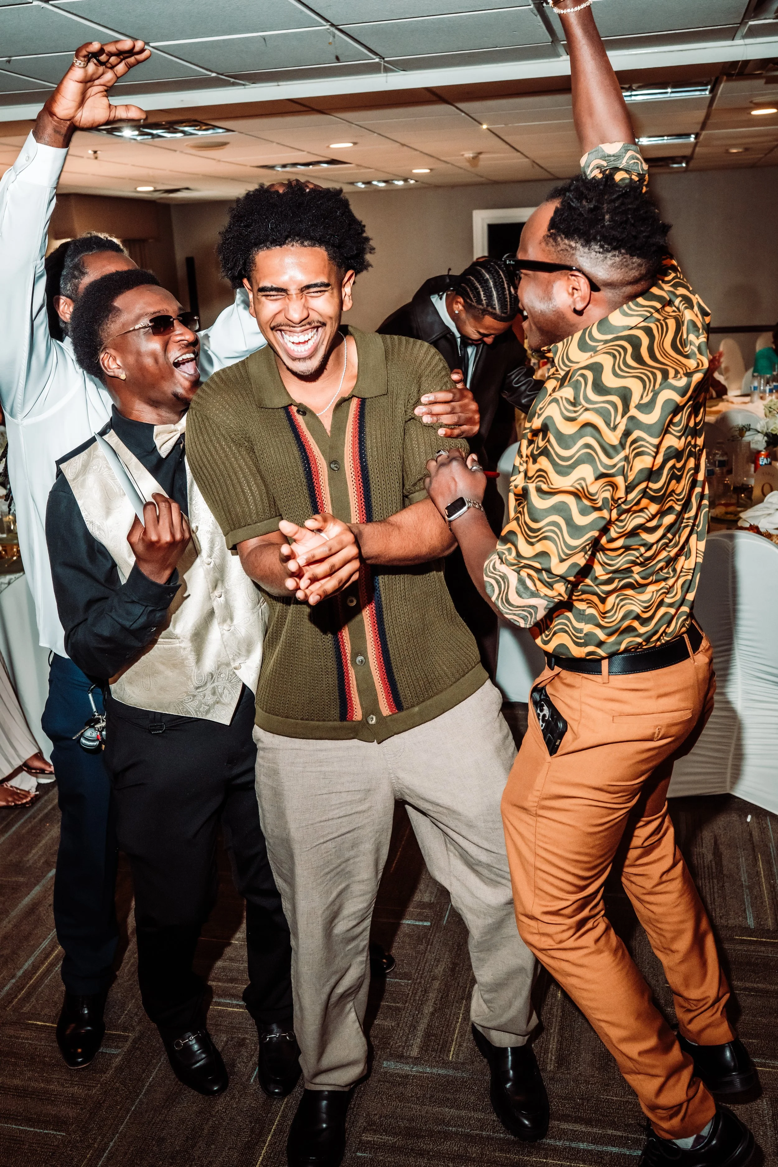 A group of six men in formal and casual attire dancing and laughing together at a celebration or party in a banquet hall.