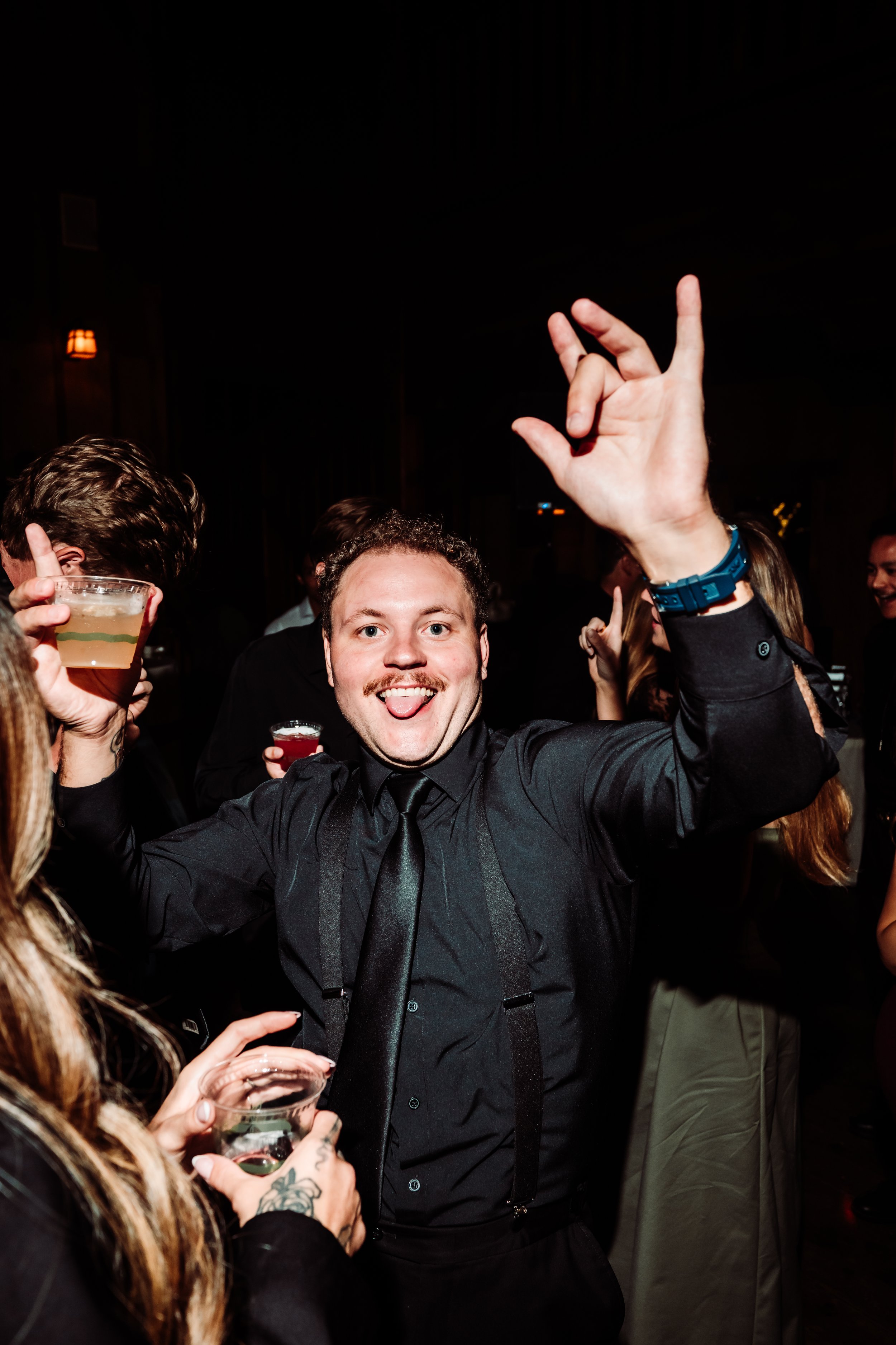 A man with short curly hair, wearing a black shirt and black tie, is smiling and sticking out his tongue at a party. He is making an 'OK' sign with his right hand and holding a drink in his left hand. The background shows other people with drinks, an