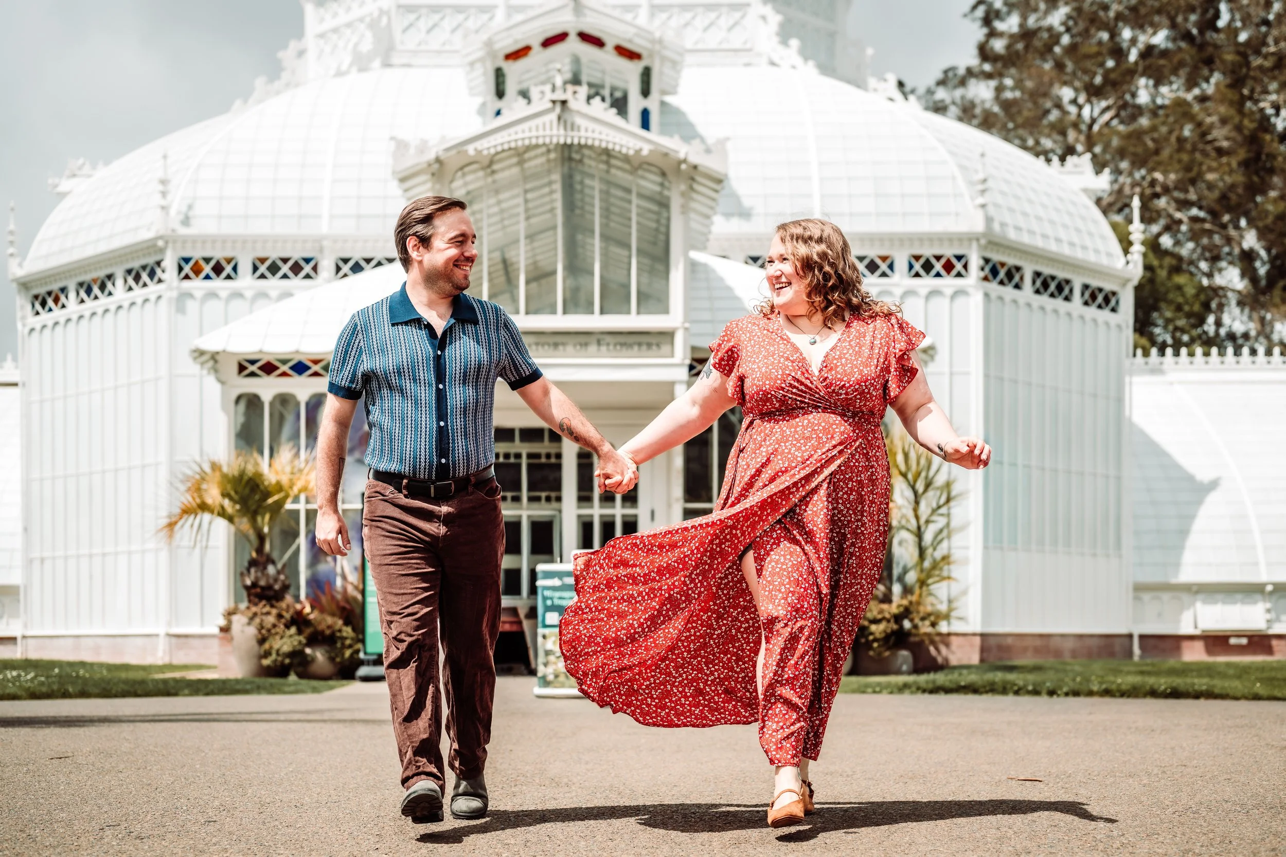 A smiling man and woman holding hands and walking outside in front of a large white Victorian house, with trees in the background. The woman is wearing a long, red floral dress and the man is in a short-sleeved, blue patterned shirt and brown pants.