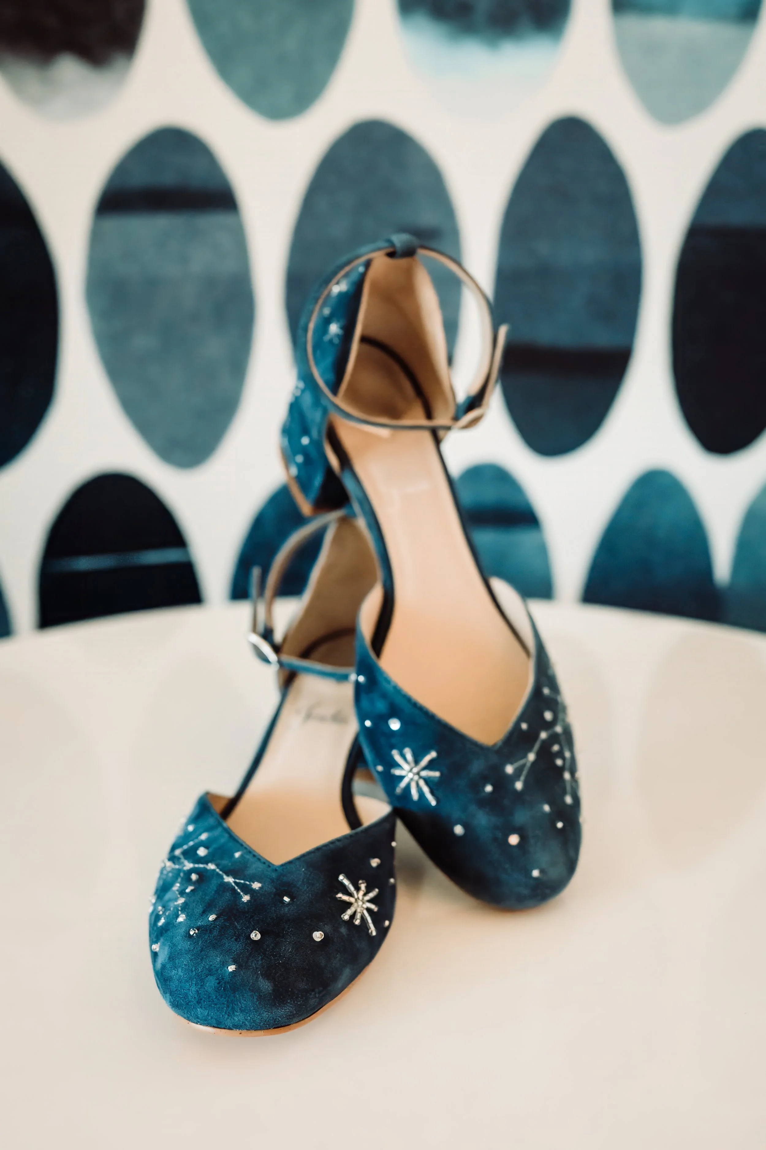 Close-up of a pair of blue high-heeled shoes with white snowflake and dot patterns, placed on a plain white surface against a background with dark oval shapes.