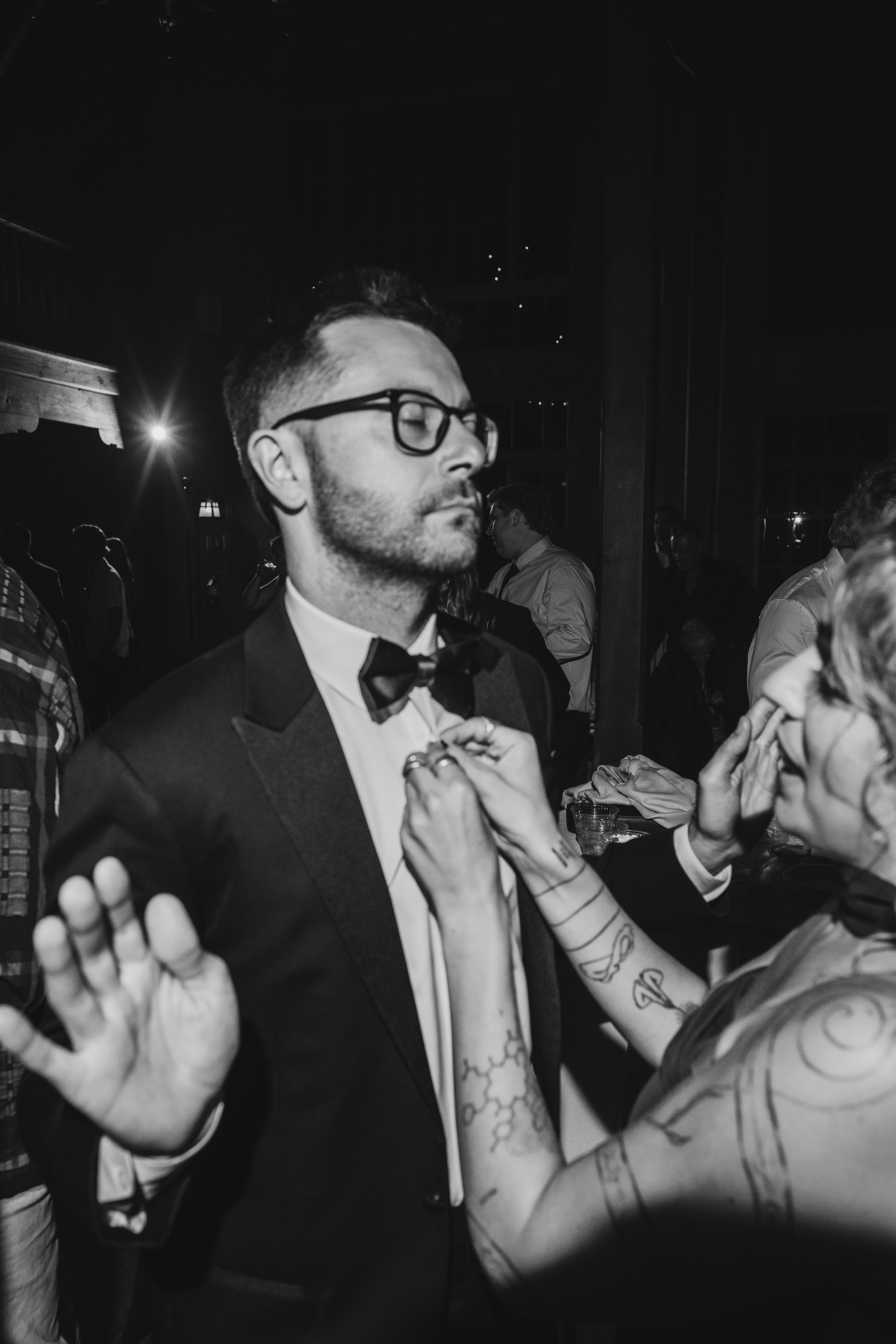 A man in a tuxedo with glasses has his eyes closed and is standing with his hands raised. Several tattooed people are reaching toward him, holding his hands and arms, in a dark, indoor setting with other people in the background.