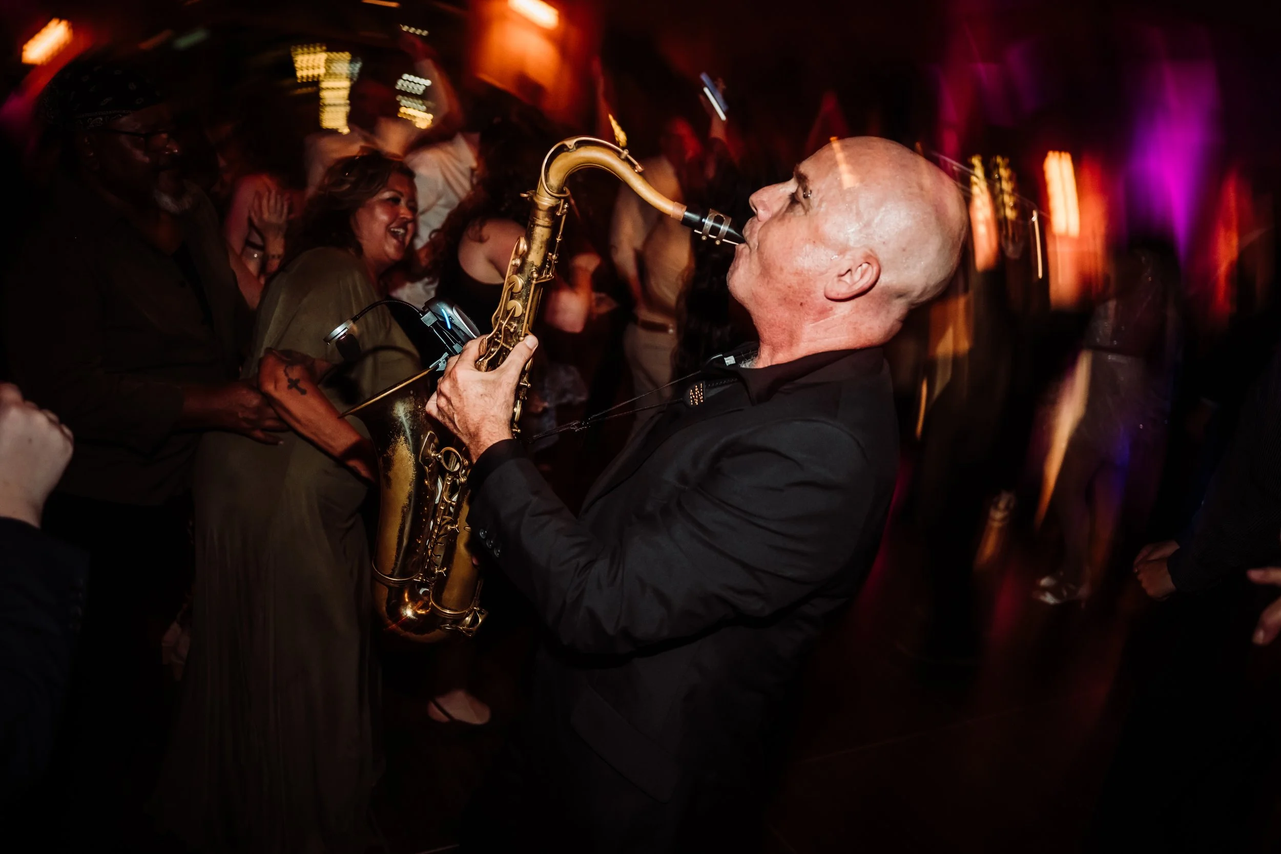 A musician plays the saxophone in a lively, colorful club scene with smiling audience members.