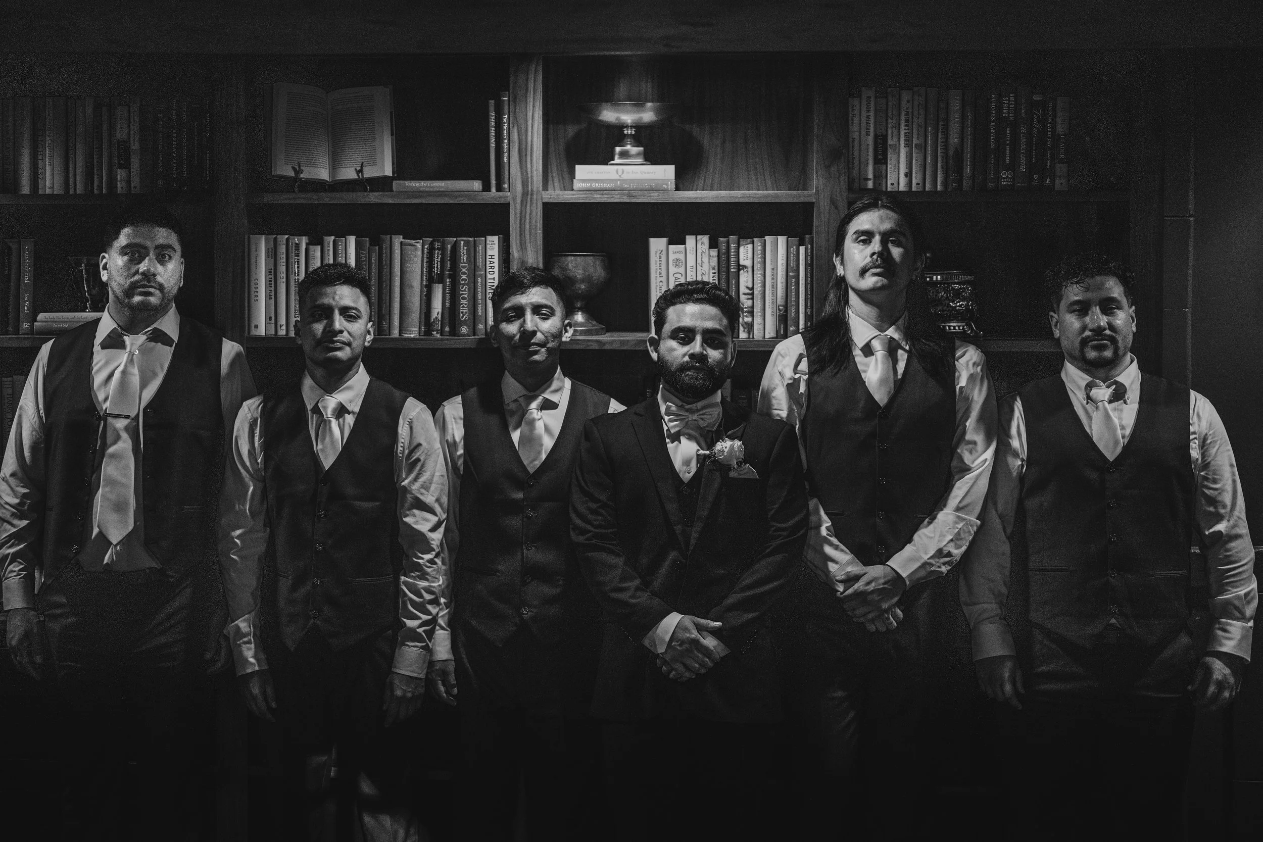 Black and white photograph of seven men dressed in formal attire, standing in front of a wooden bookshelf filled with books.