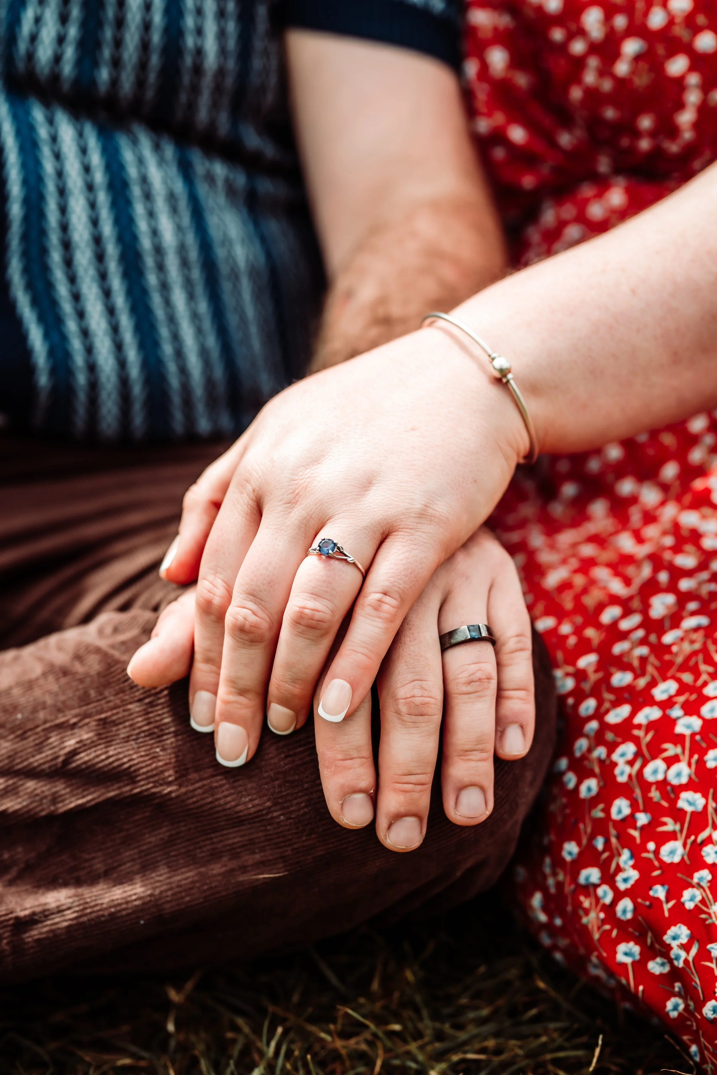 Close-up of hands resting on each other, one hand wearing a silver bracelet, another with a silver ring with a blue gemstone, on a background of a person wearing a red floral dress and another person in a blue sweater and brown corduroy pants.