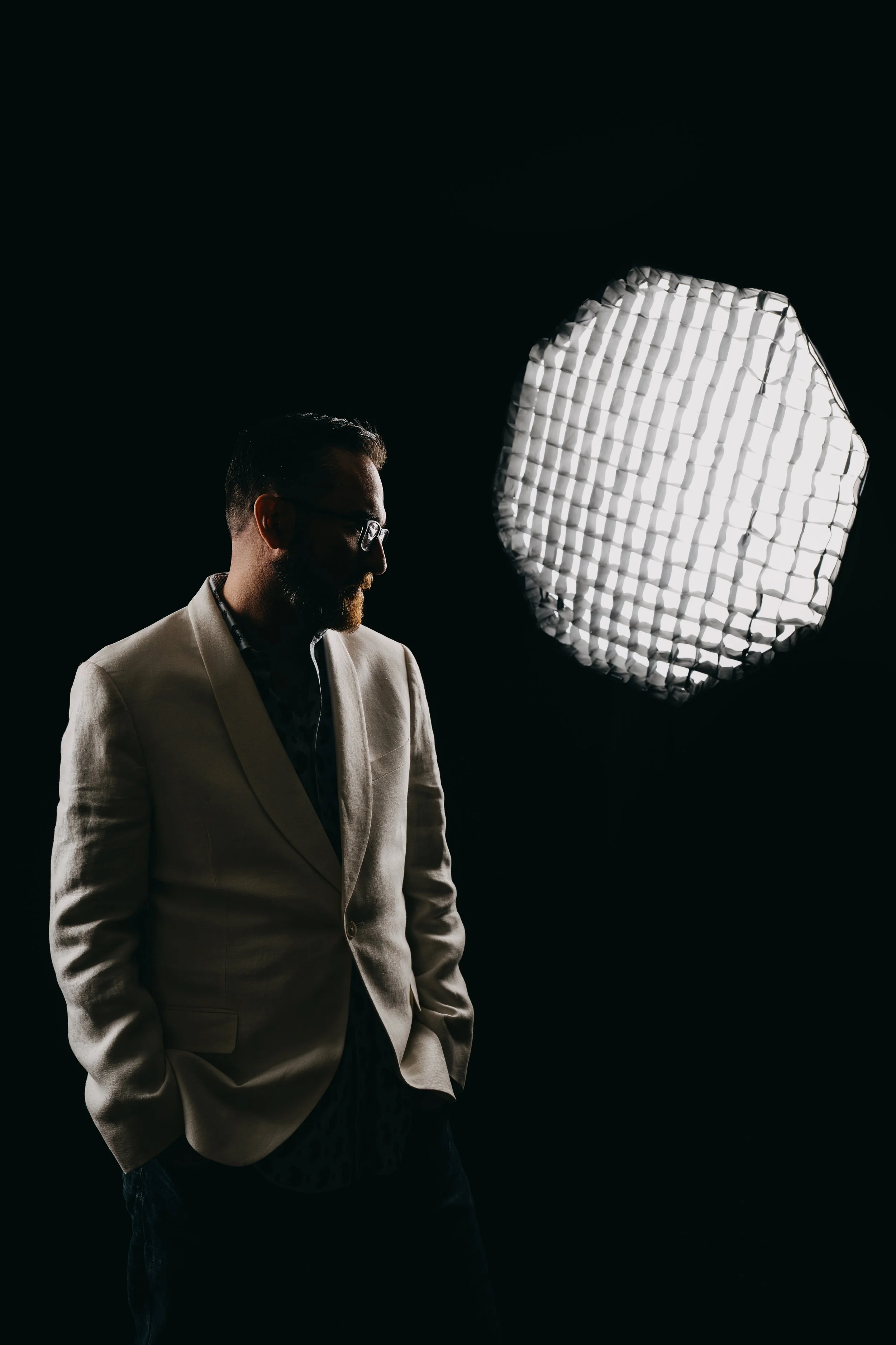 A man wearing glasses and a light-colored blazer stands in front of a large studio light with grid diffusion, against a dark background.