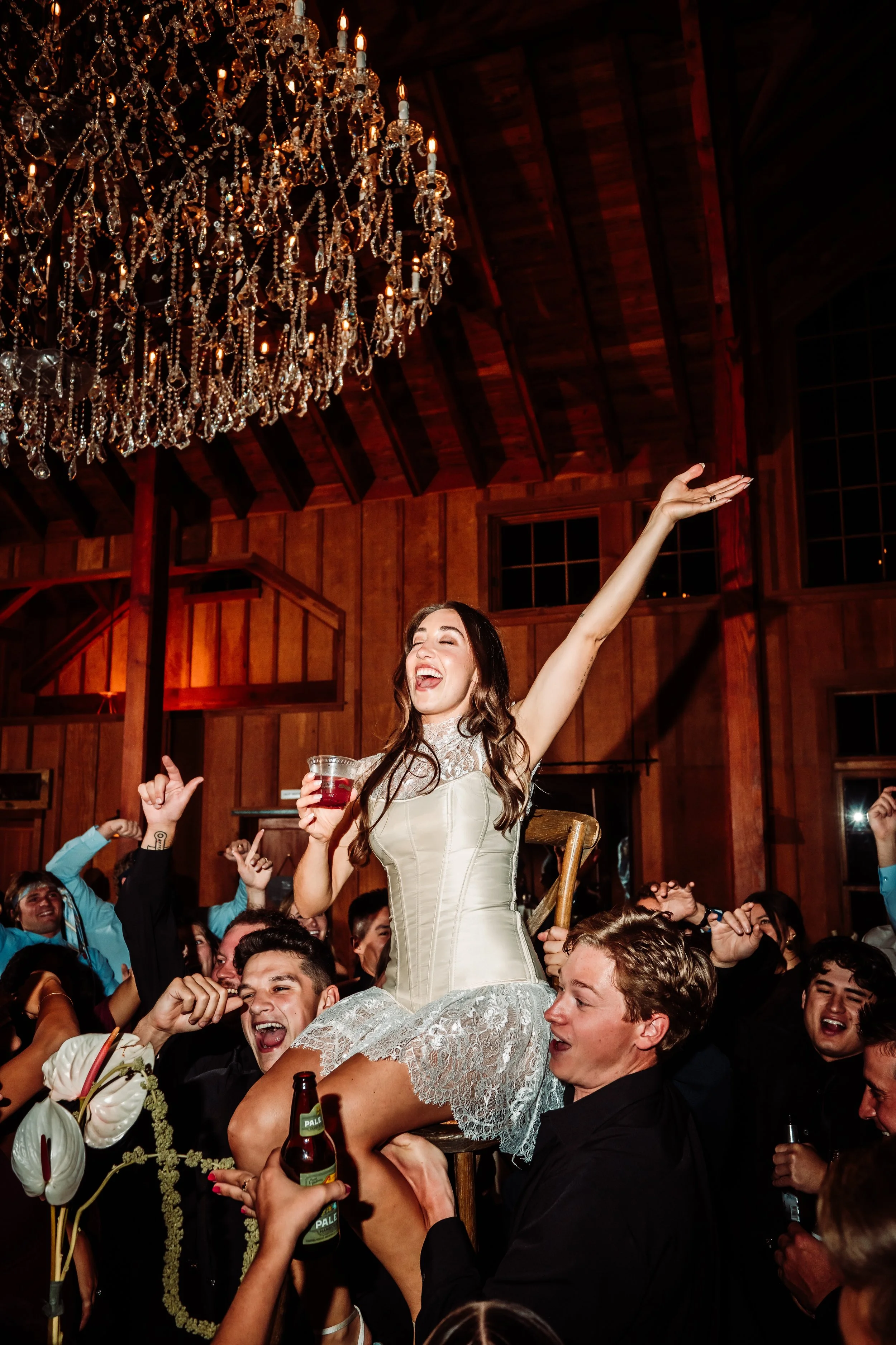 A woman in a sleeveless wedding dress being lifted on the shoulders of a large group of people at a celebration, with a chandelier hanging from the ceiling and wood-paneled walls in the background.