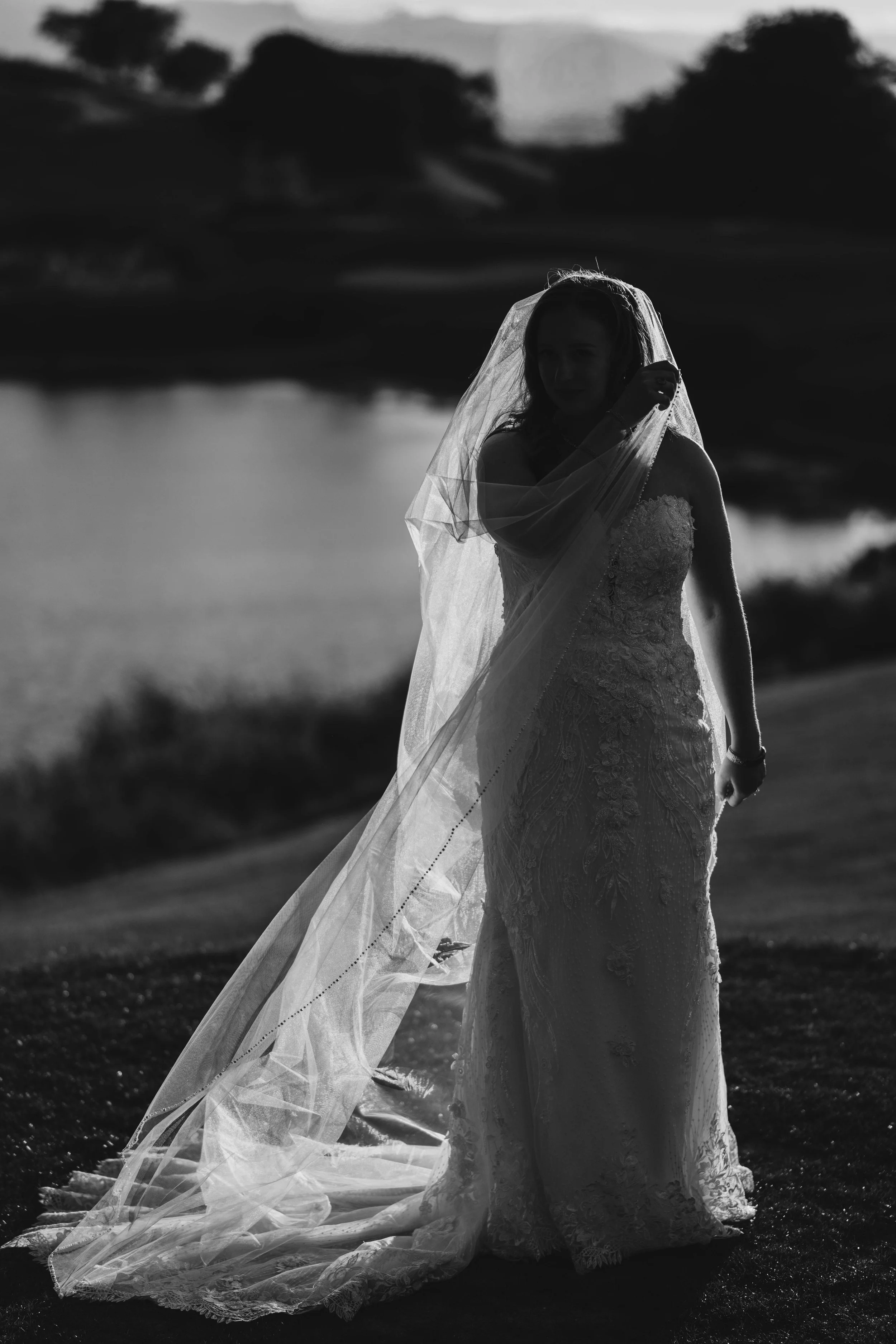 A bride in a lace wedding gown and veil stands outdoors by a body of water, with a scenic background of trees and hills, captured in black and white.