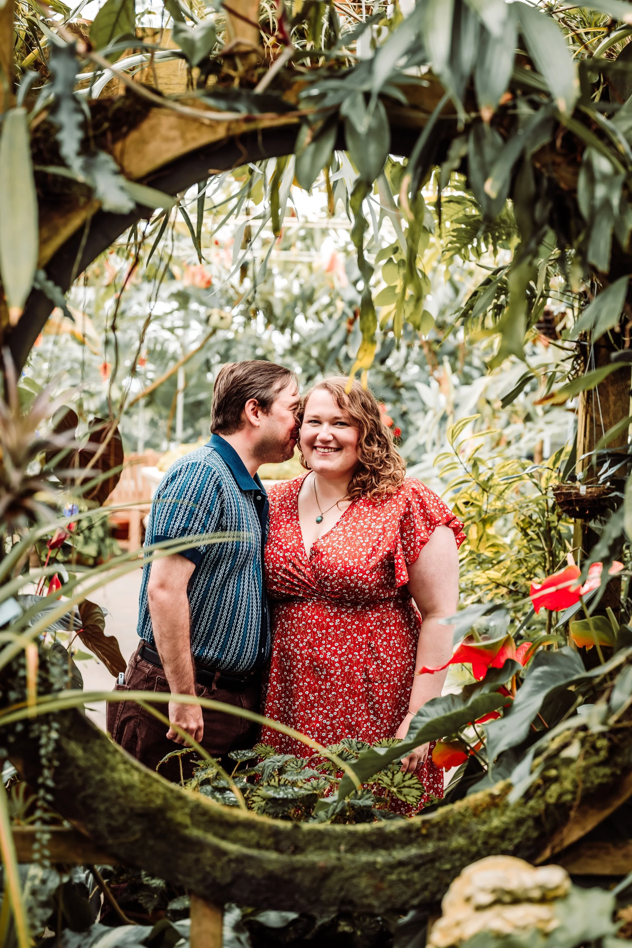 A young man and woman standing close together in a green, leafy garden, with the man whispering into the woman's ear and both smiling; the image is framed by tree branches and plants.