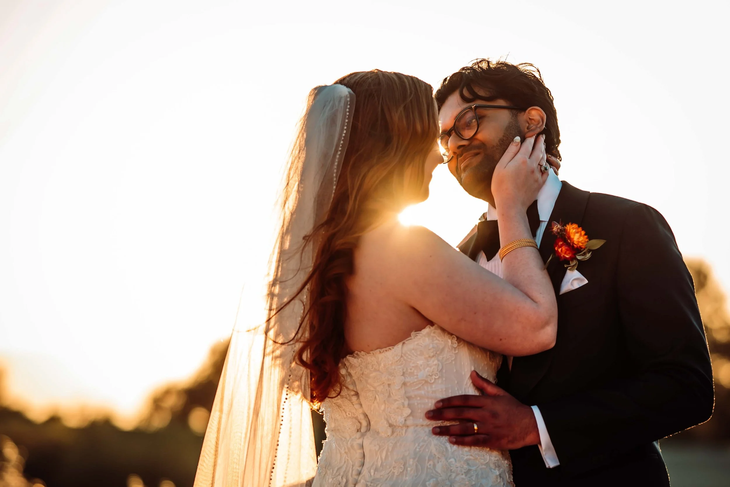 A bride and groom embrace during sunset, with their faces close together, smiling and touching each other's necks, outside.