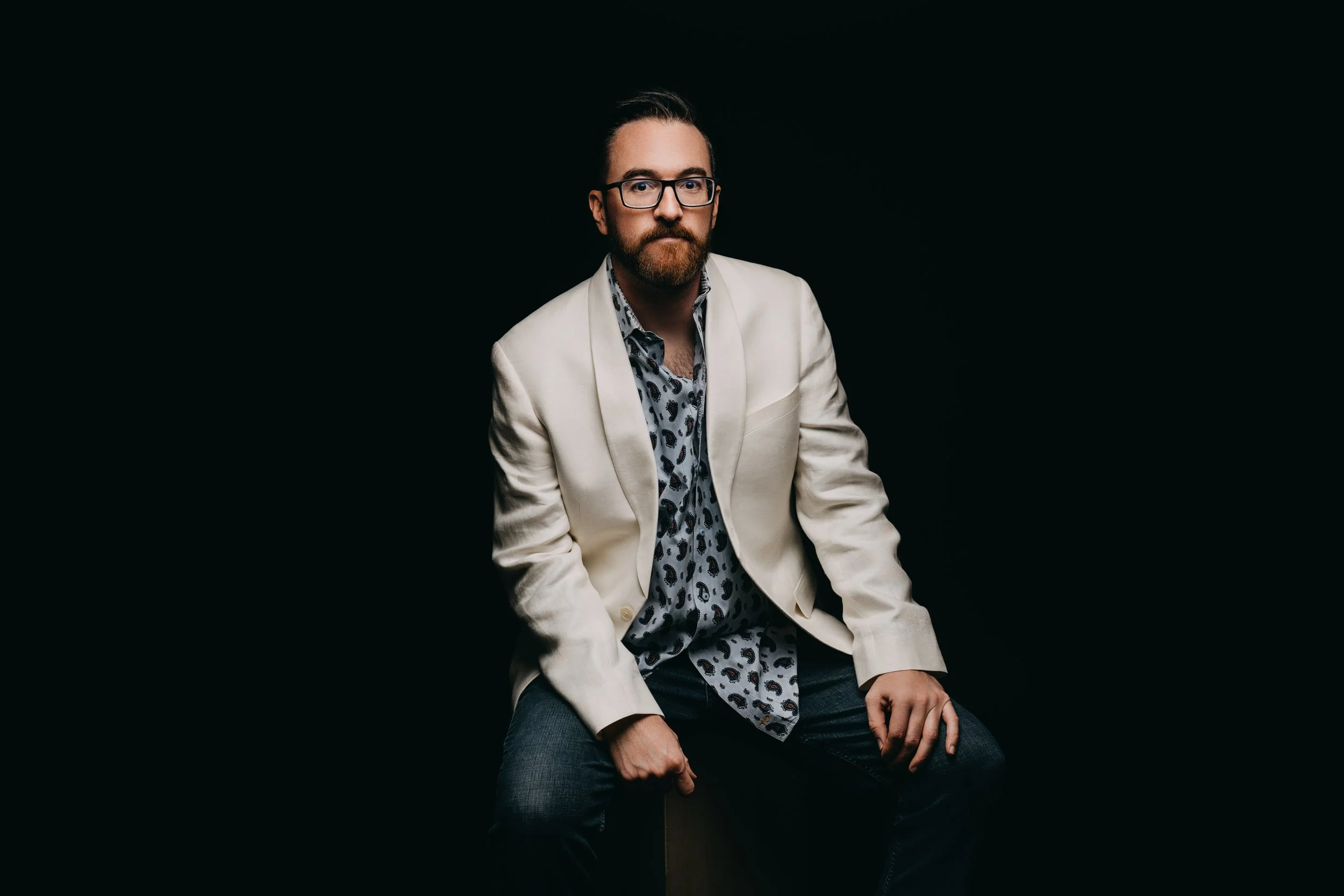 A man with glasses, a beard, and mustache wearing a light-colored blazer, a patterned shirt, and dark jeans, seated against a dark background.