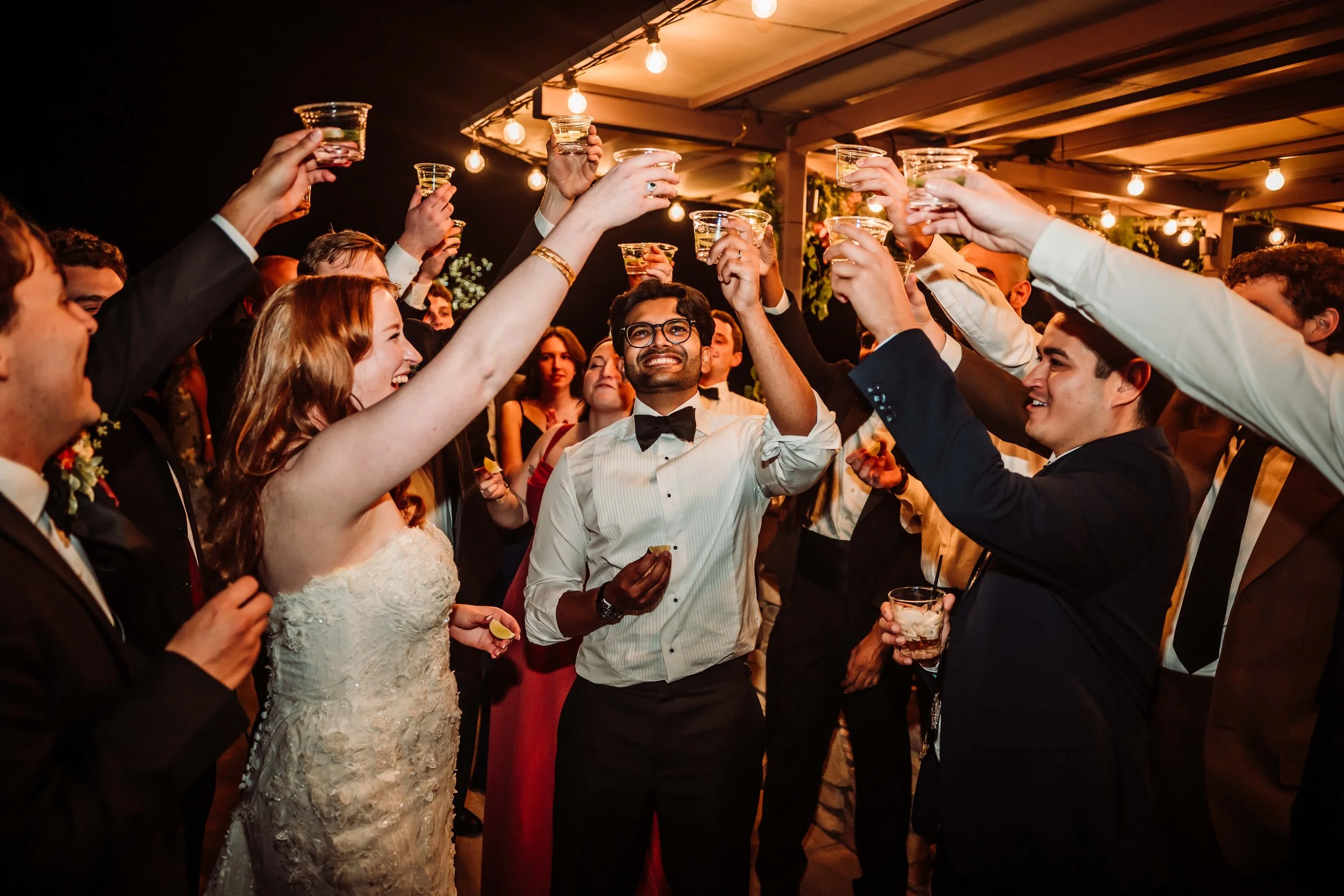A group of people at a party raising glasses for a toast. The scene is festive with warm lighting and a decorated outdoor or semi-outdoor area.