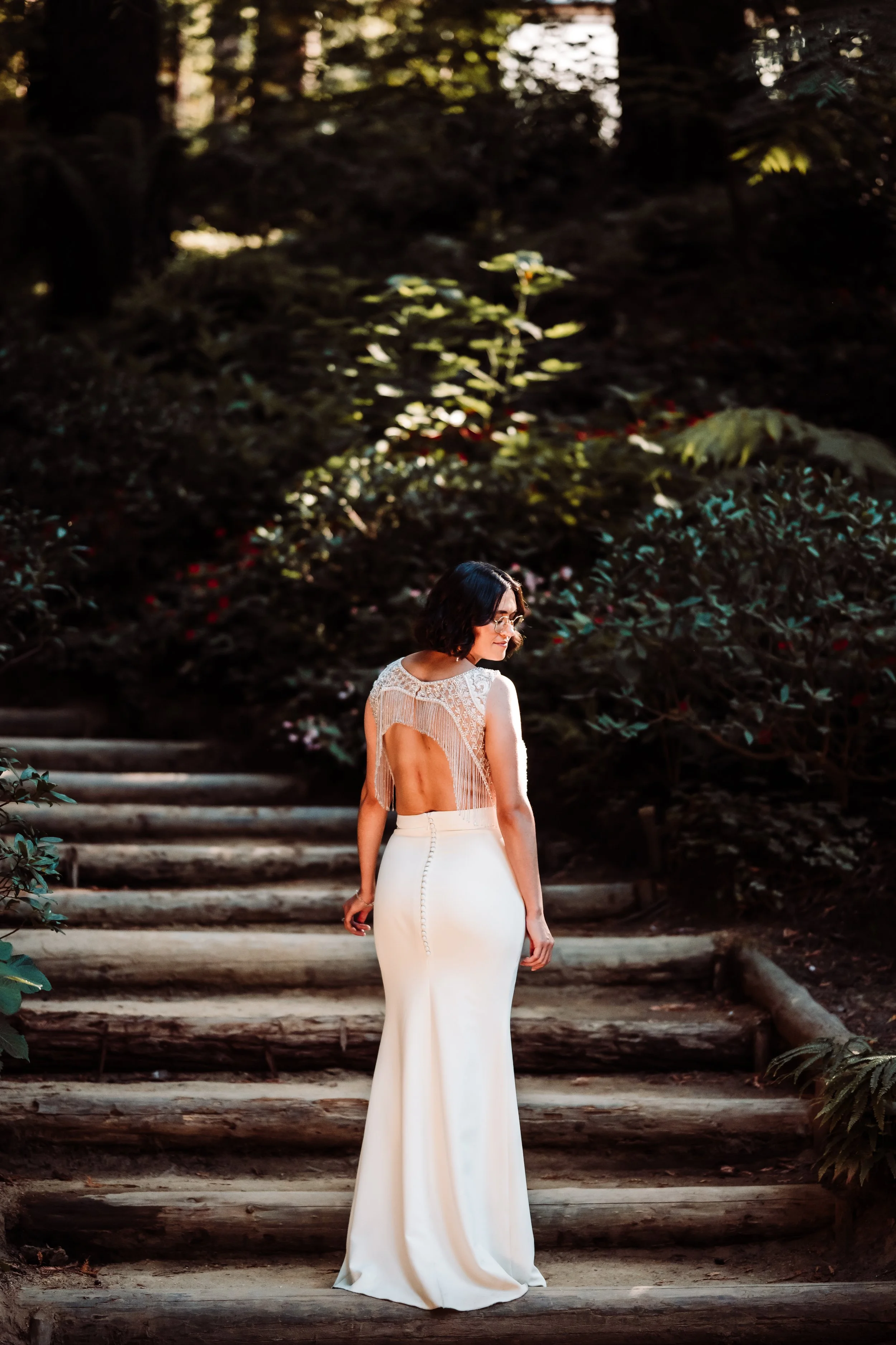 A woman with short dark hair and glasses standing on wooden steps outdoors in a white, backless, floor-length dress with lace details, surrounded by green foliage.