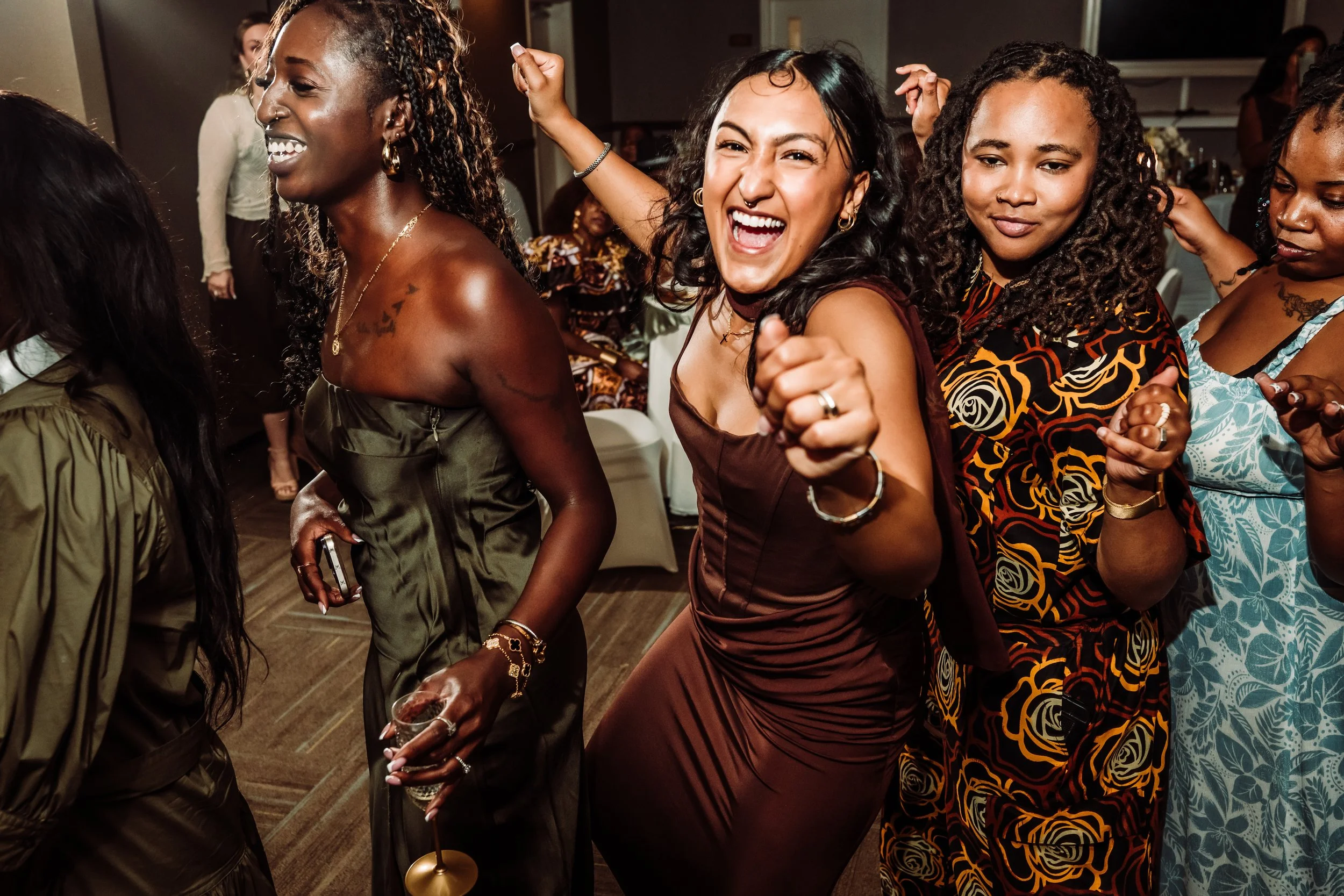 Four women enjoying a celebration, dancing and smiling, dressed in elegant outfits at an indoor party.