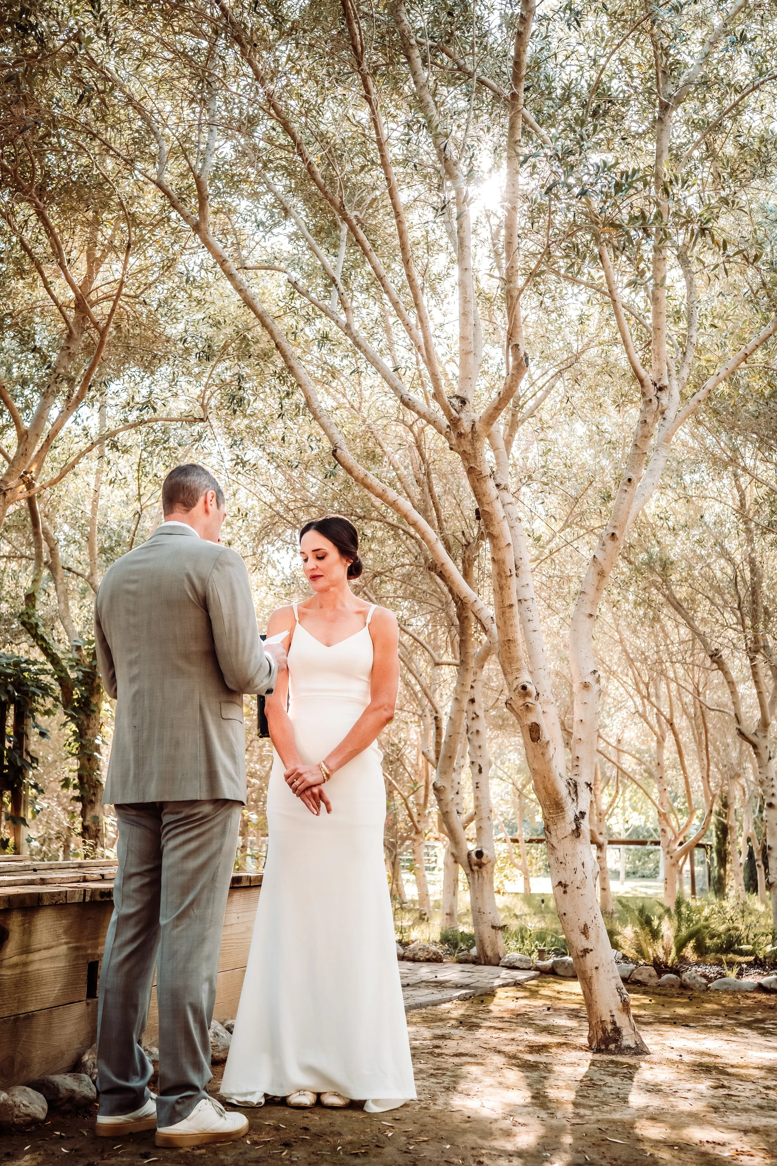 A couple dressed in wedding attire standing outdoors among trees during daytime. The man is in a gray suit; the woman is in a white wedding gown, looking at each other.