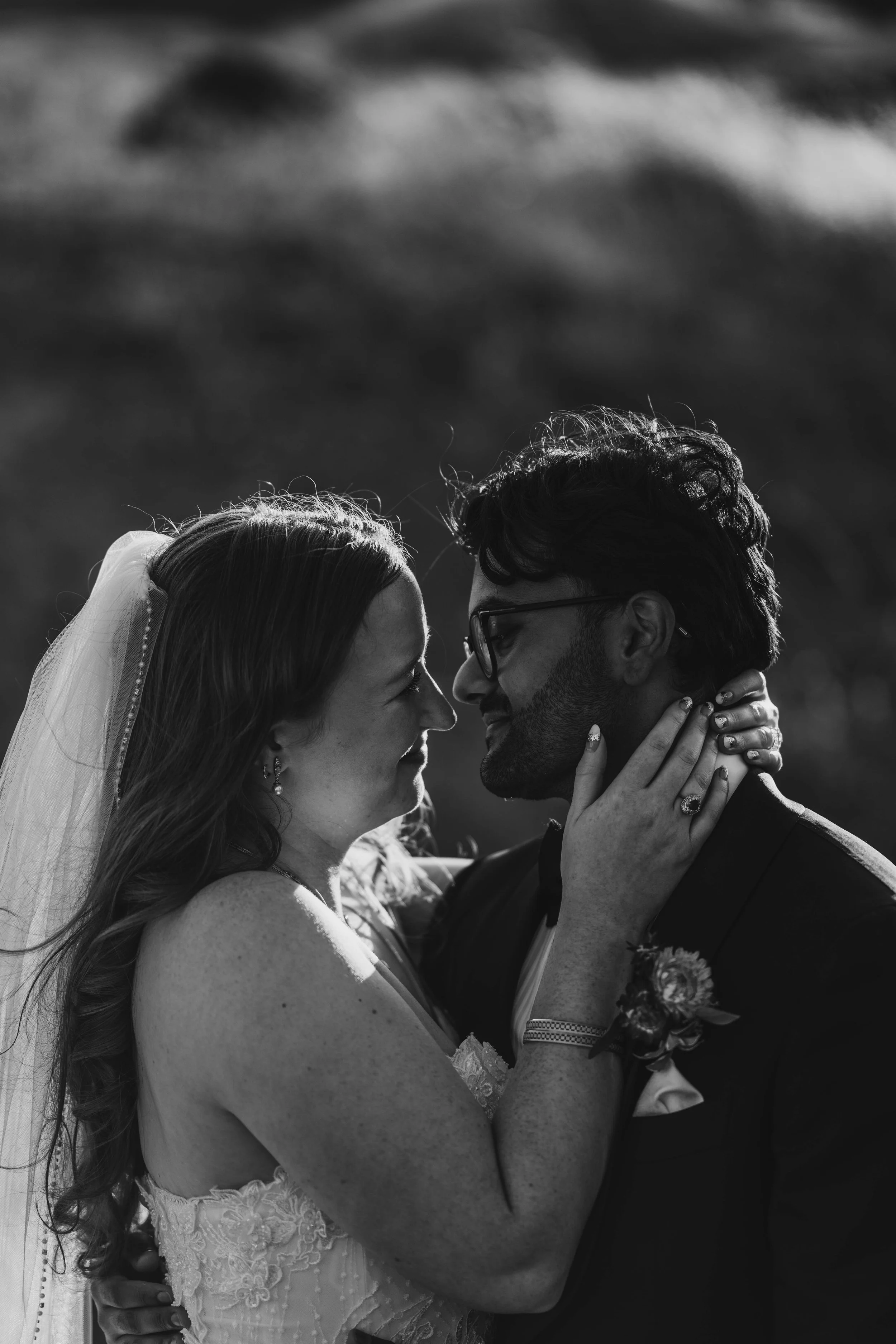 A black and white photo of a bride and groom embracing and about to kiss outdoors. The bride is wearing a lace wedding dress and veil, and the groom is wearing glasses and a dark suit with a boutonnière.