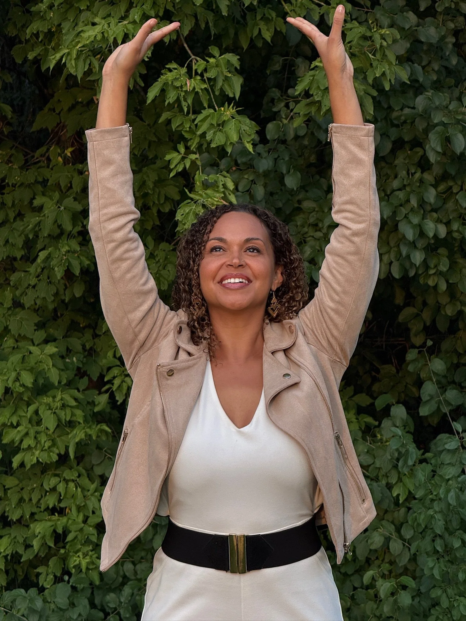 Dr. Ihotu Ali smiles and raises her arms above her head against a leafy green background, wearing a beige jacket over a white top and a black belt.