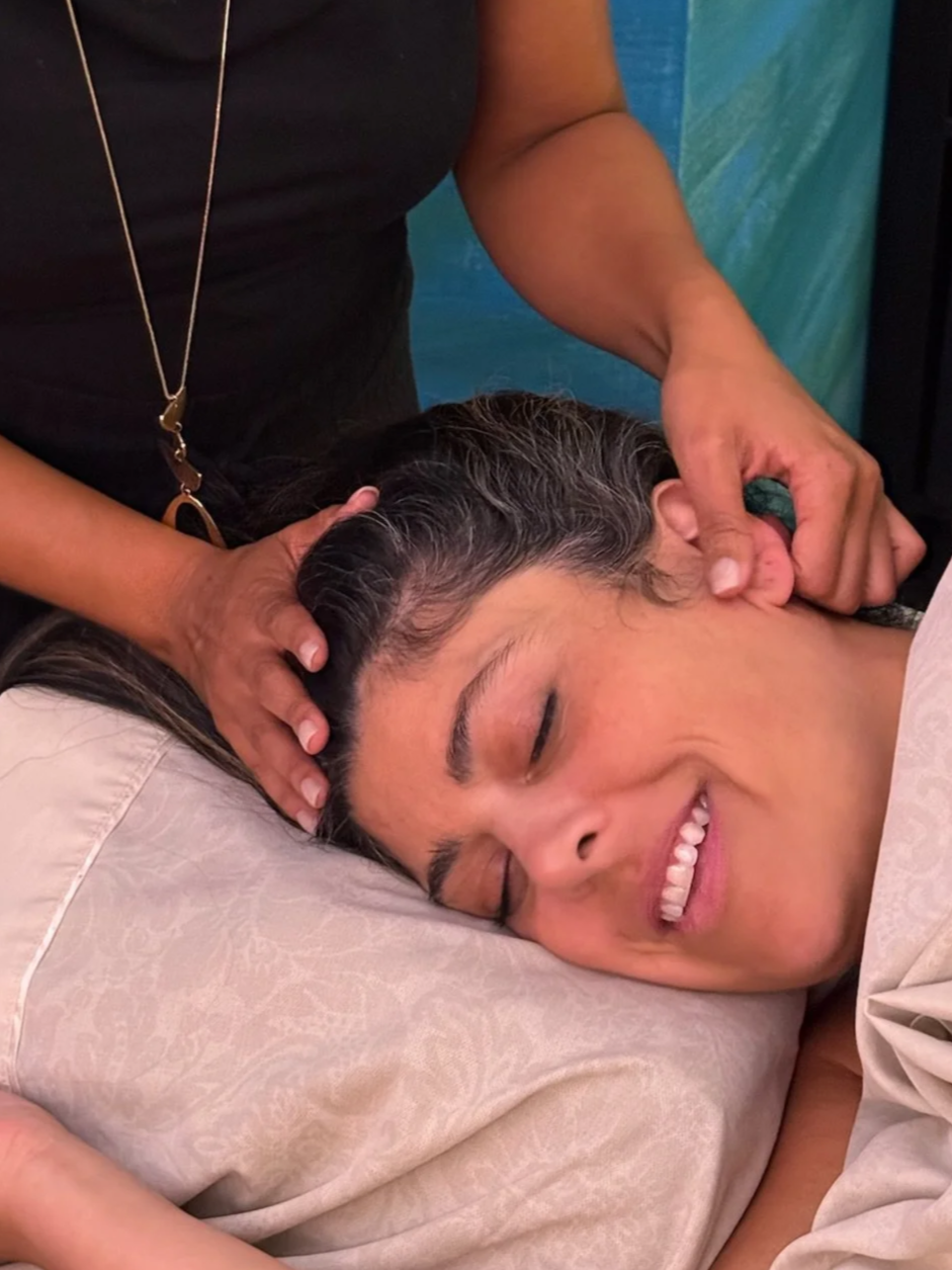 A woman lying with her eyes closed, smiling, receiving a massage on her head and neck from Dr. Ihotu Ali.