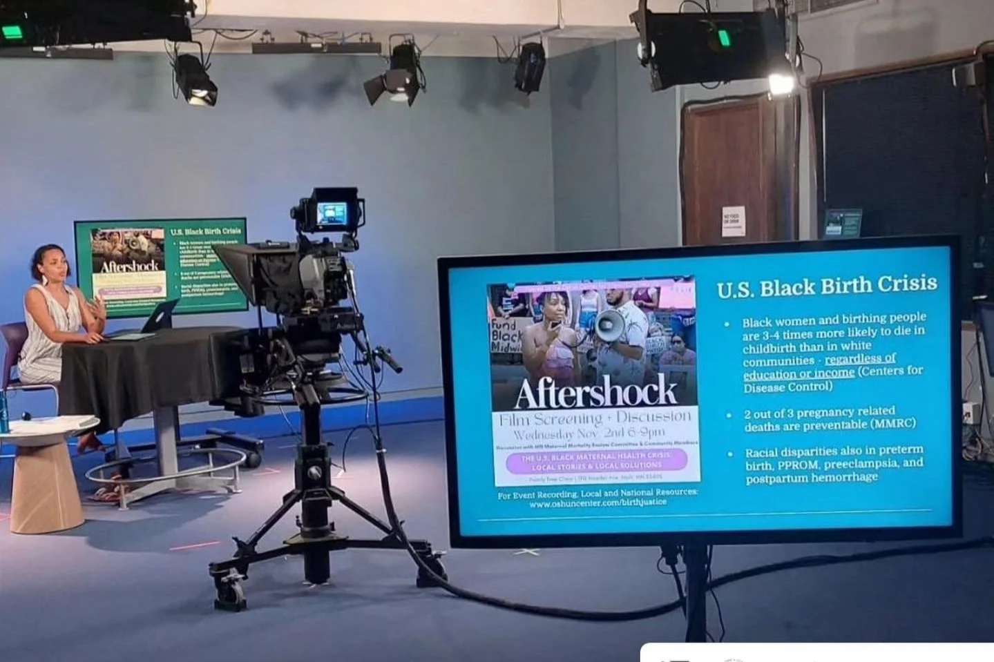 A news studio with Dr. Ihotu Ali sitting at a desk with a laptop, a large camera on a tripod, and a large screen displaying information about a film screening and discussion on the U.S. Black birth crisis.