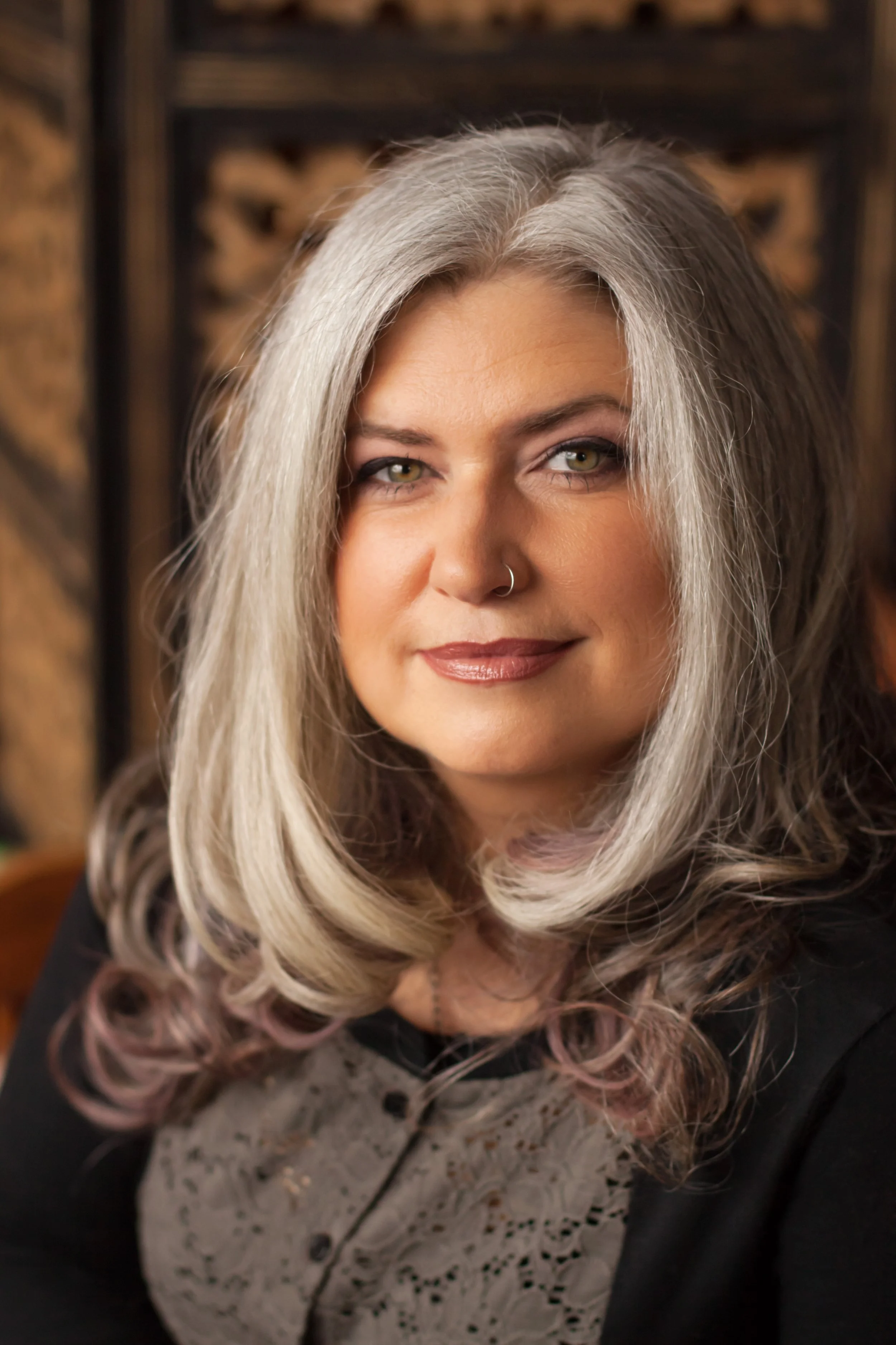 Close-up of Dr. Michele Renee with silver-gray hair, light skin, wearing a nose ring, and a patterned shirt. She is smiling slightly and has green eyes. Background is an ornate wooden carved decorative panel.