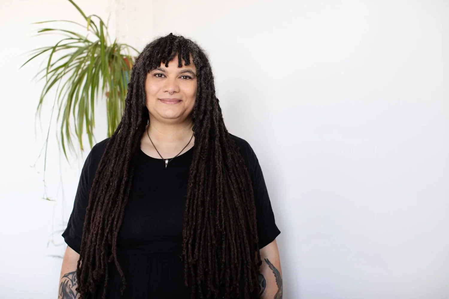 Nathalie of Anansi Acupuncture,  with long dreadlocks, wearing a black shirt and necklaces, standing in front of a white wall with a potted plant in the background.