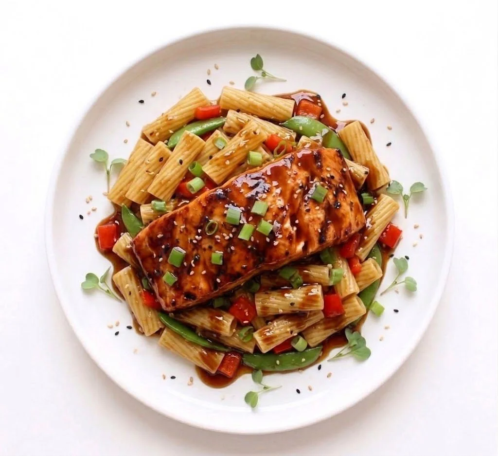 Grilled salmon fillet topped with sauce, served with pasta, diced red and green bell peppers, scallions, and black sesame seeds on a white plate.