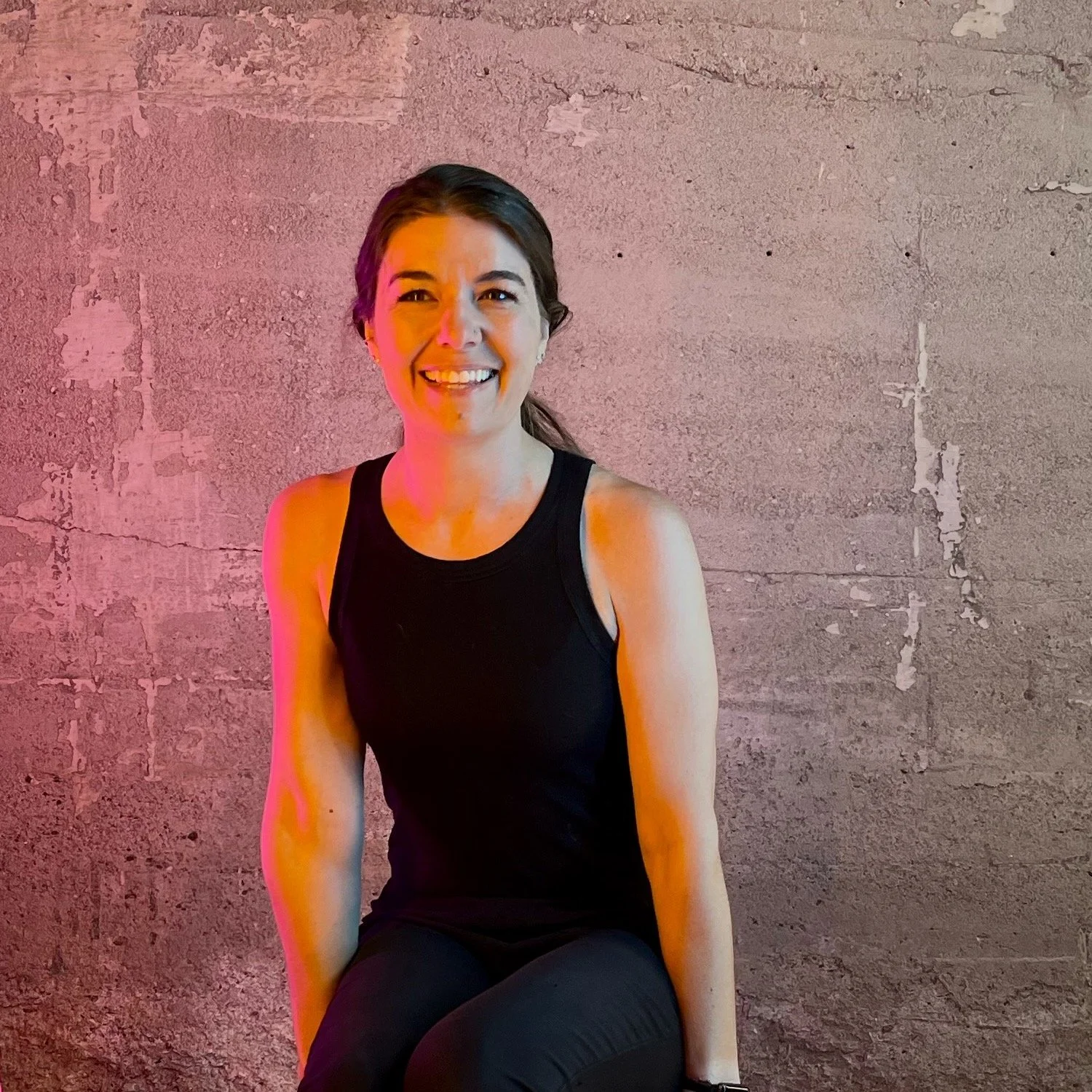 A woman in a black sleeveless top sitting against a textured pink wall, smiling at the camera.