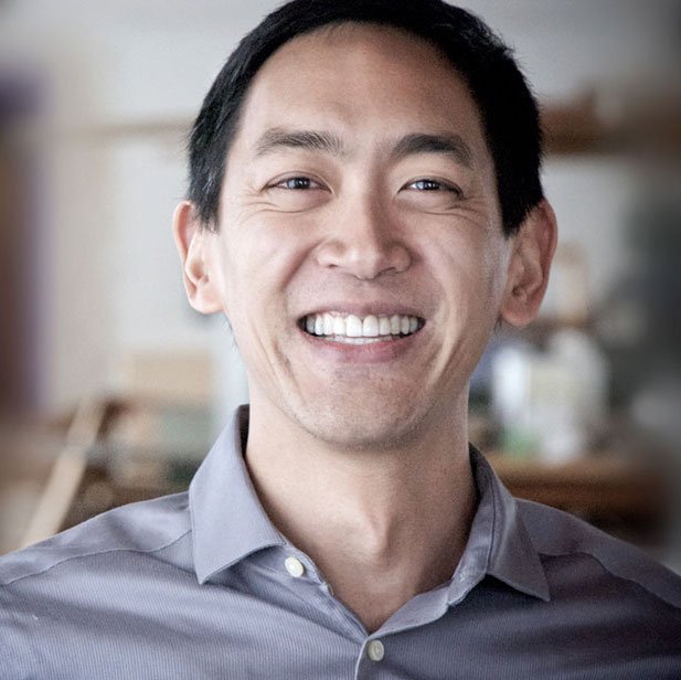 A smiling man with short black hair wearing a light gray collared shirt in an indoor setting.