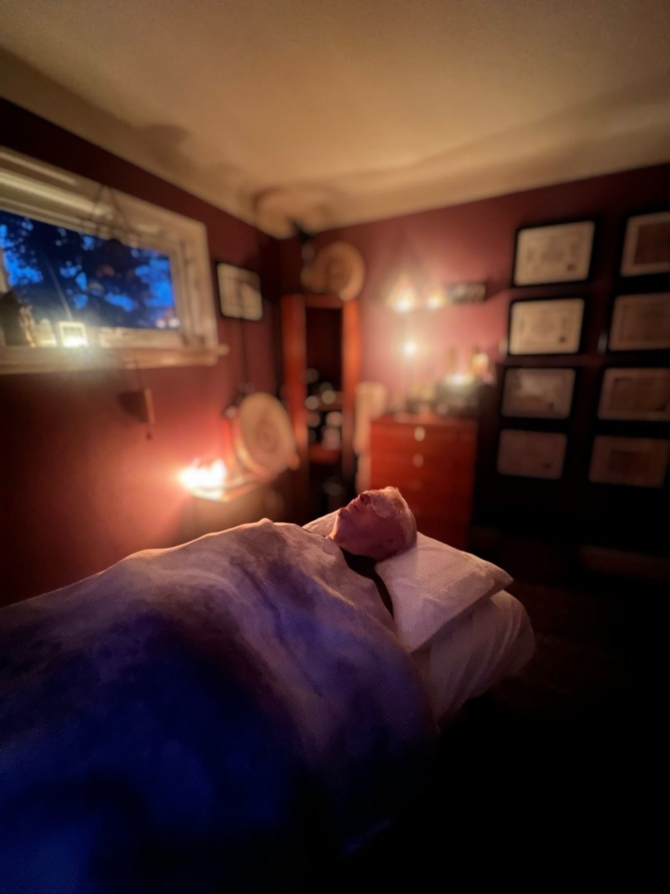 A person receiving a reiki healing session in a room with dim lighting, a window showing a blue evening sky, and framed pictures on the wall.