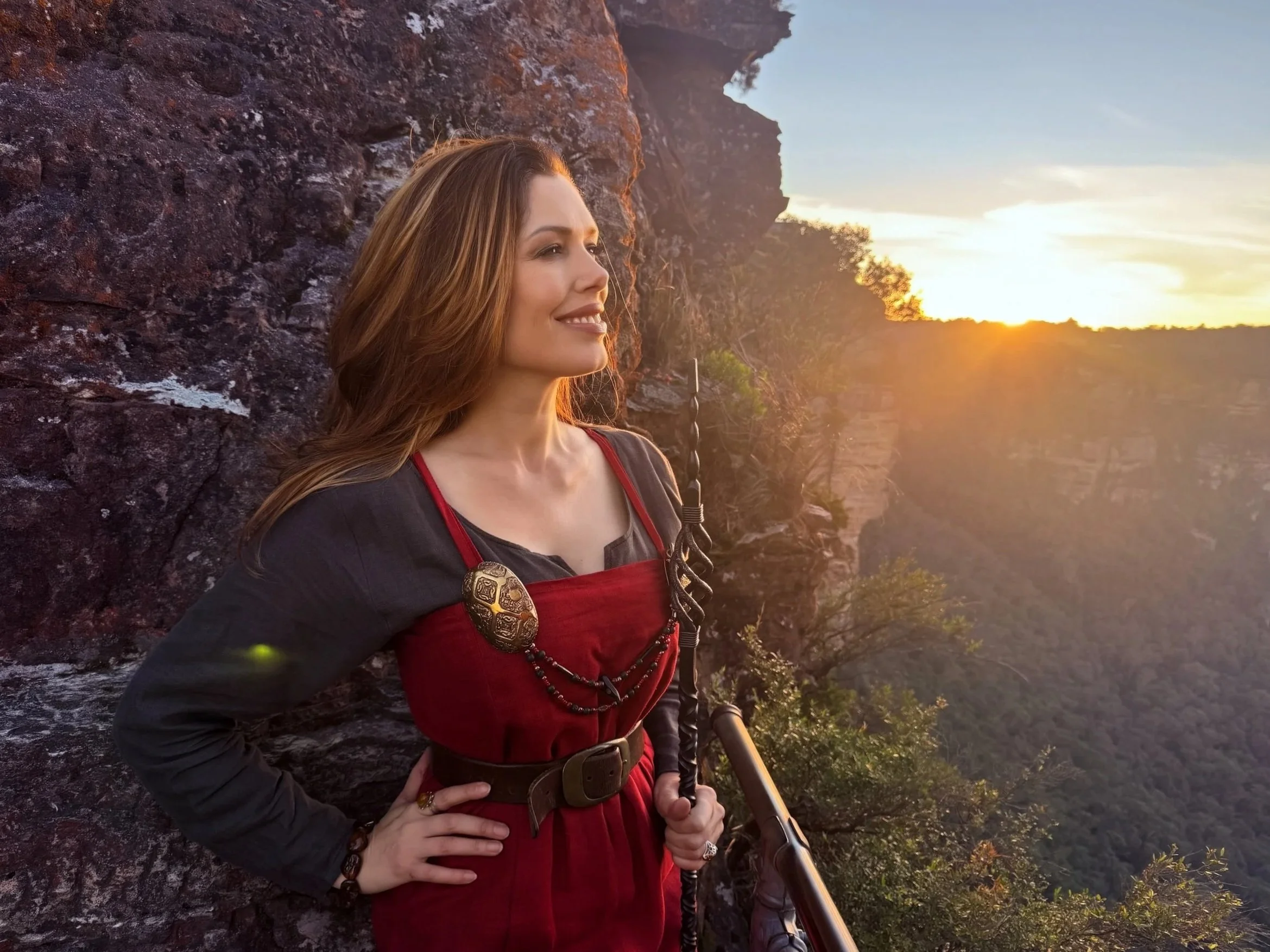 Rae Moss, a woman with long hair, dressed in a red traditional Norse Viking woman outfit with jewelry, standing outdoors at sunset near rocky terrain and trees, holding a Seidr staff.