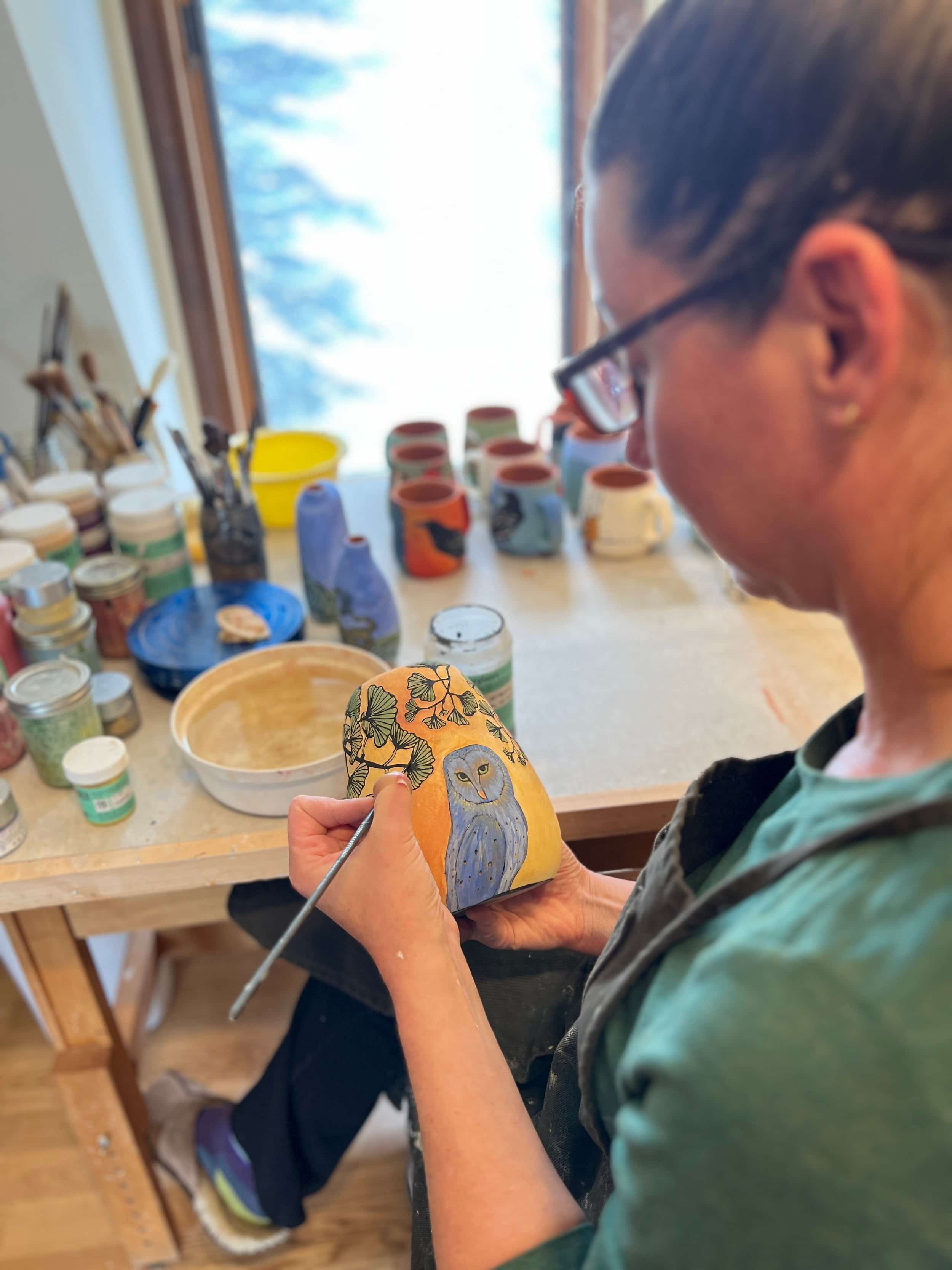 The ceramic artist, Ishara, is painting ginkgo leaves and a barn owl on a vase.
