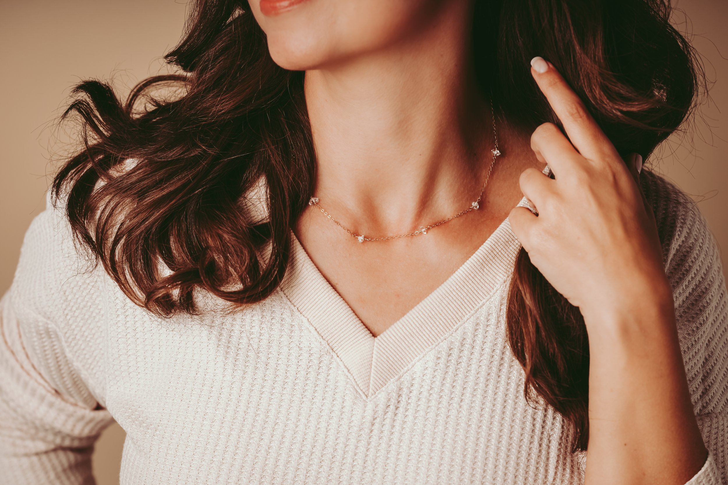 Close-up of a woman wearing a delicate necklace and ring, with dark wavy hair and a white textured V-neck sweater, touching her neck and ear.