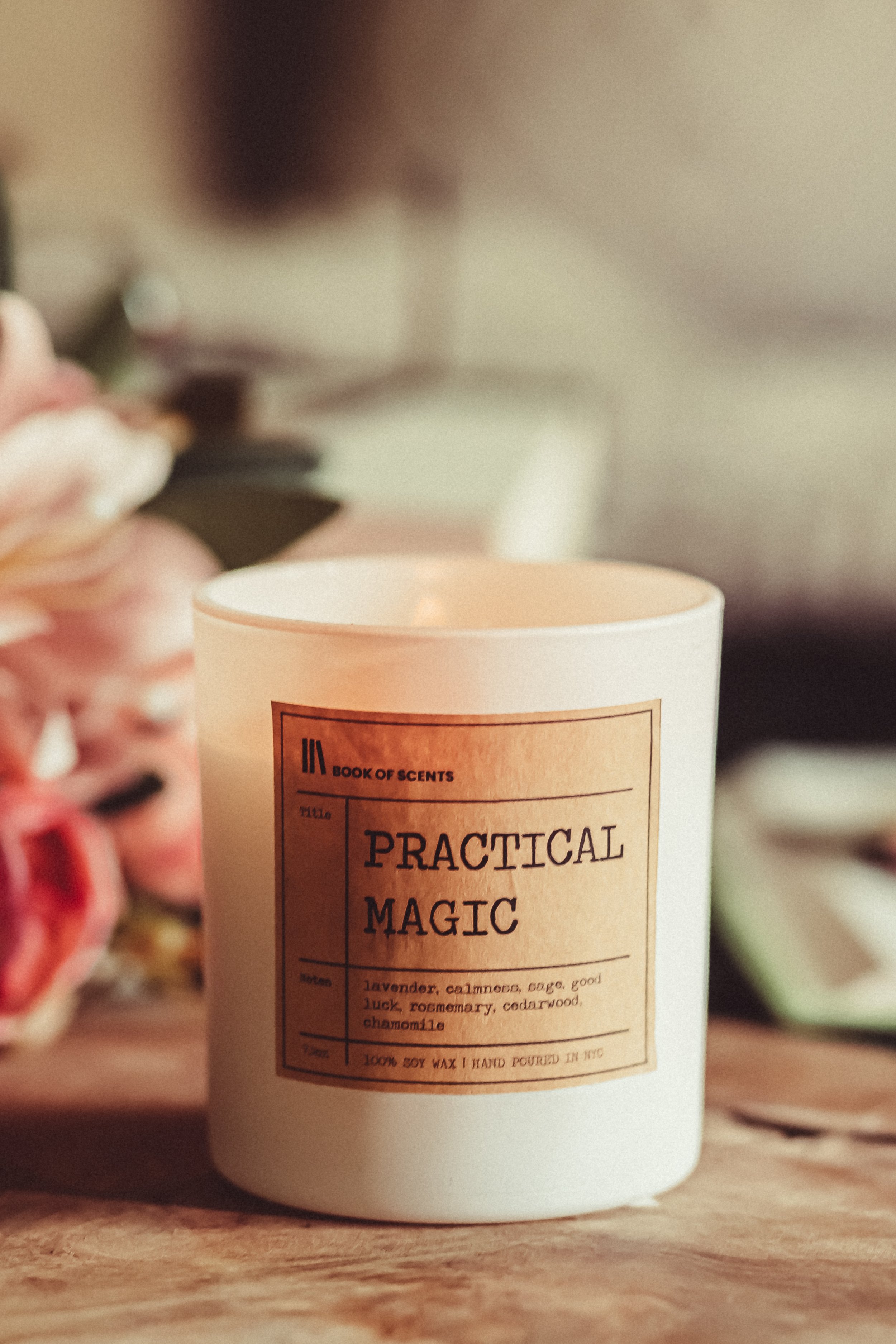 A white candle with a label reading 'Practical Magic' on a wooden surface, with pink flowers and other objects blurred in the background.