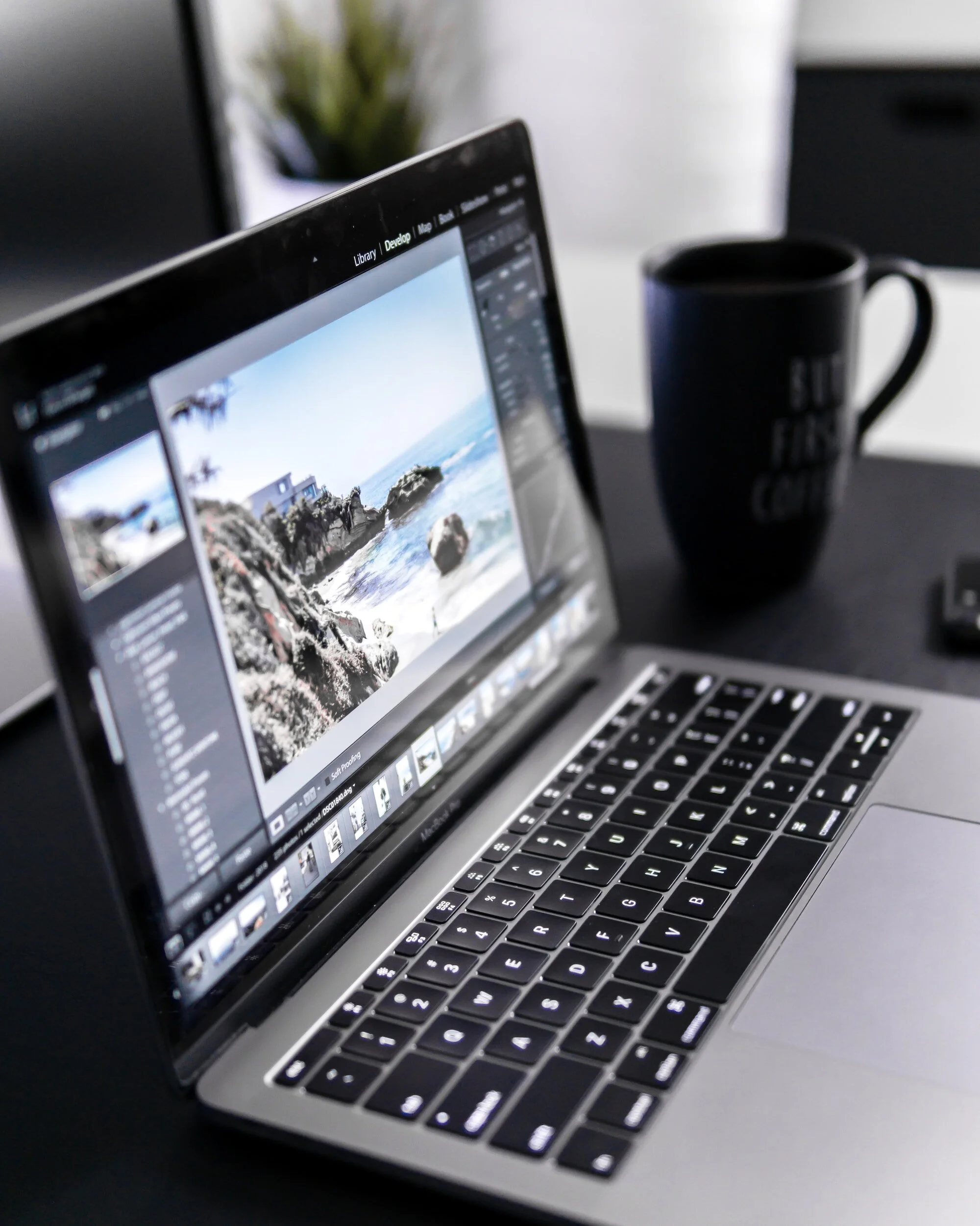 A laptop on a desk displaying a photo editing software with a beach scene, next to a black coffee mug.