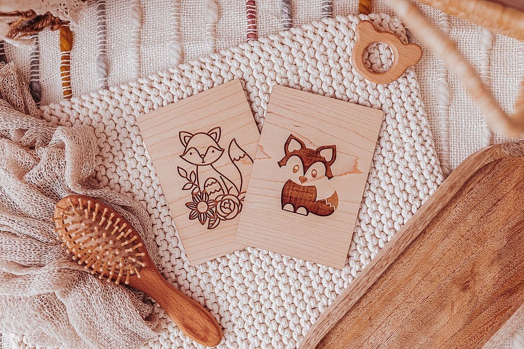 Wooden cards with engraved animal illustrations of a fox and a raccoon, a wooden hairbrush, a baby teething ring, and a textured blanket on a woven surface.