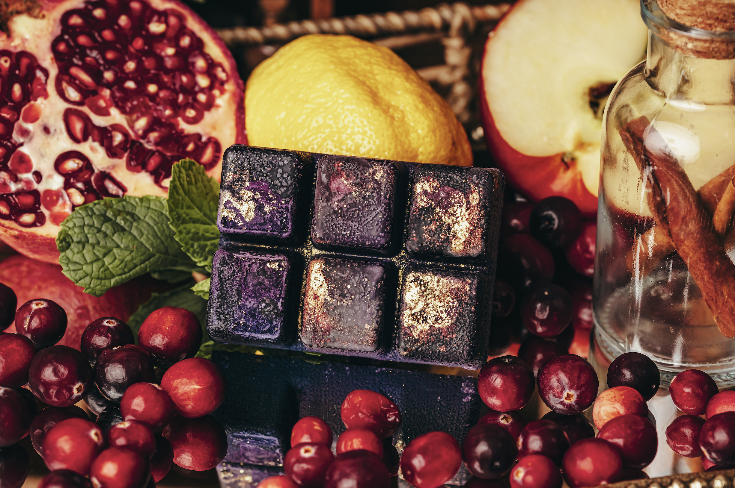 Assorted fruits, including pomegranate, lemon, apple, and cranberries, surrounding a black chocolate cube with purple and gold accents, along with a jar of water with cinnamon sticks and a piece of ginger.