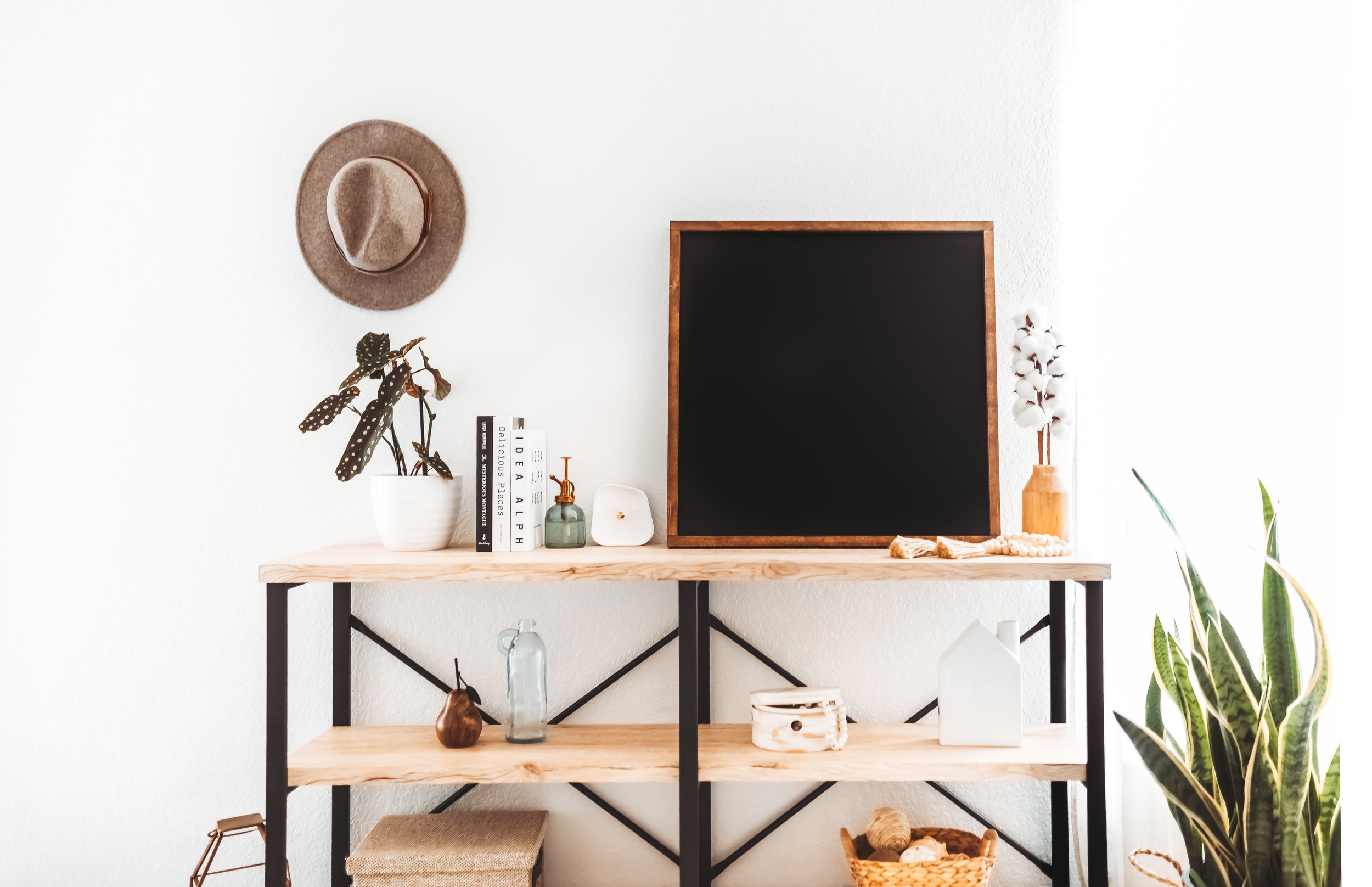Wood shelf against a white wall decorated with books, a small plant, a lantern, a white house-shaped decor, a small basket, and a vase of cotton stems. A hat hangs on the wall, and there is a potted plant to the right.