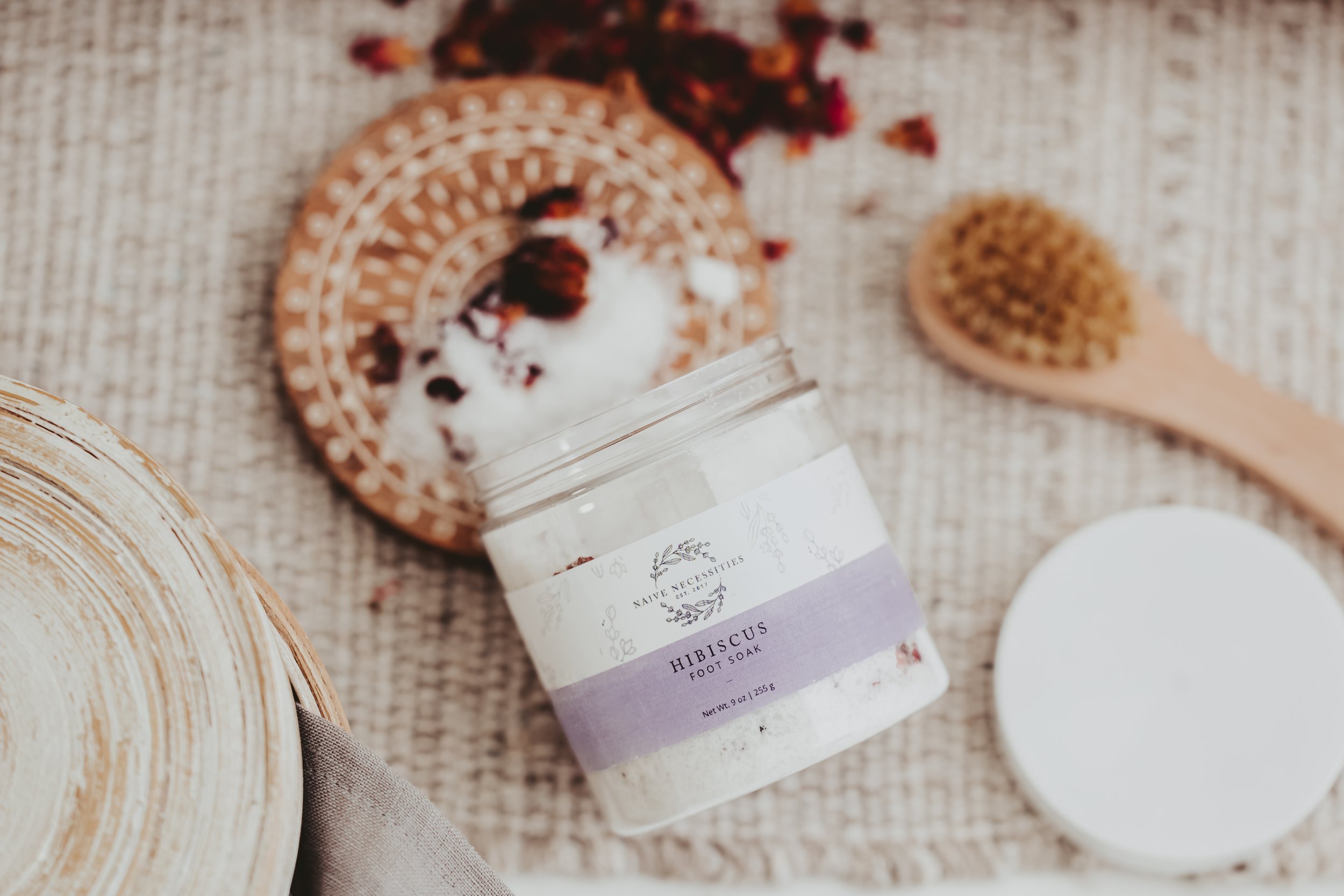 Open jar of Hibiscus foot soak surrounded by a wooden brush, a decorative woven tray, and a cloth on a textured beige surface.