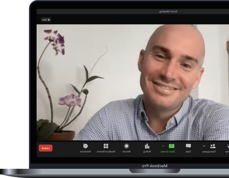 Man smiling in a virtual meeting, with a purple orchid in a pot on his left side and a white wall background.