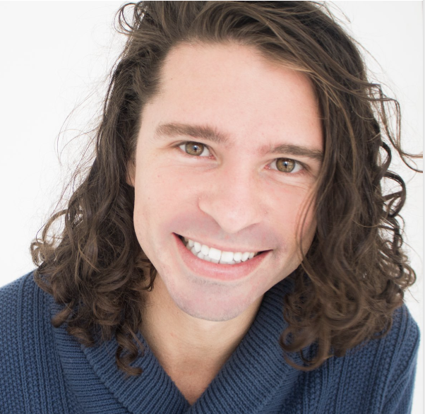 Smiling person with long, curly hair wearing a blue sweater.